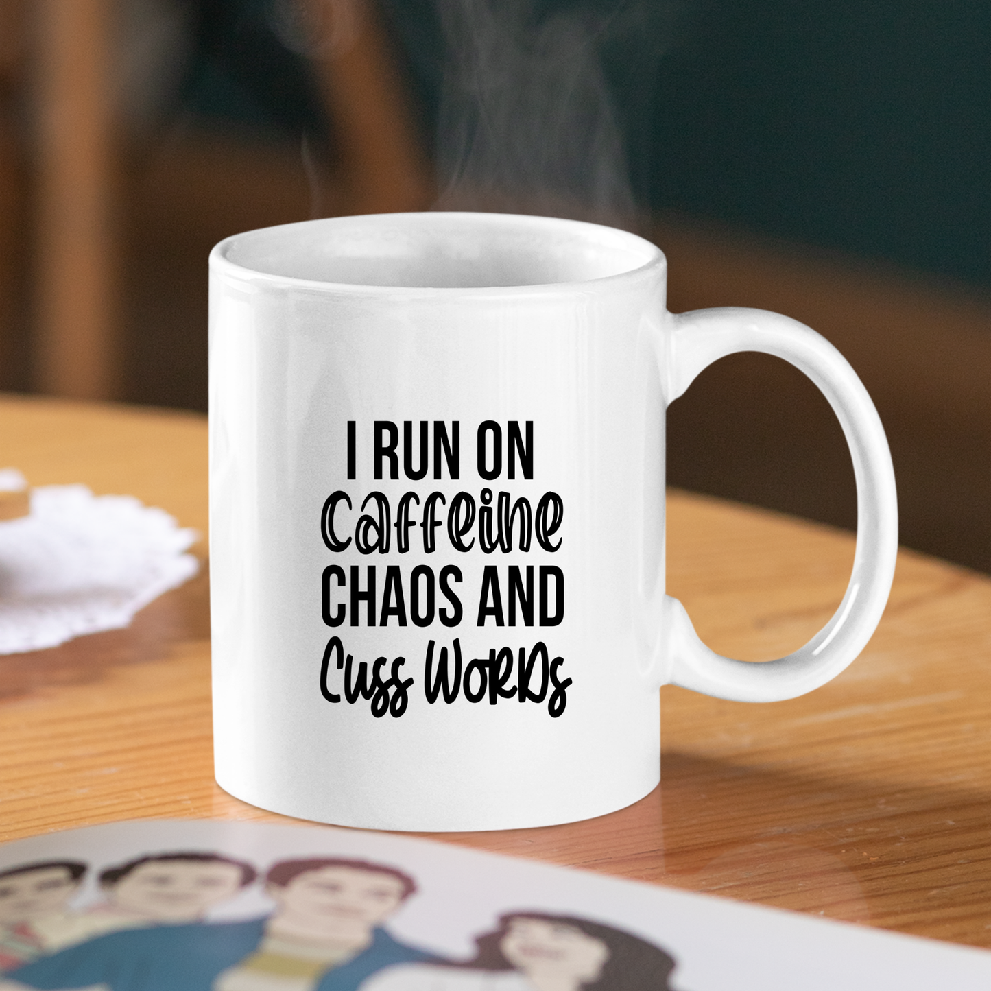 The Caffeine Chaos Cuss Words Mug | Funny Coffee Gift sits steaming on a wooden table with bold black text: “I RUN ON CAFFEINE, CHAOS AND CUSS WORDS,” while a nearby cartoon family photo adds a touch of charm to the scene.