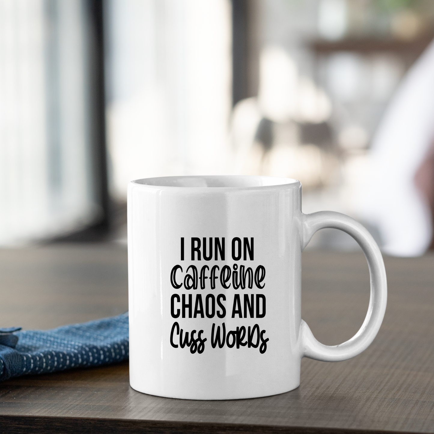 The Caffeine Chaos Cuss Words Mug | Funny Coffee Gift sits on a wooden table, bold text reading "I run on caffeine, chaos and cuss words." A folded blue cloth is nearby, with a softly blurred background creating a cozy vibe.