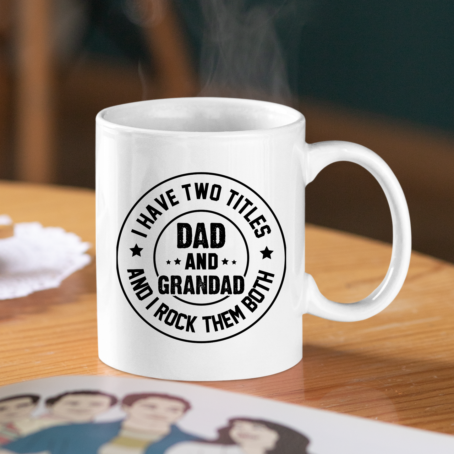 A white ceramic coffee mug on a table features the words, "I have two titles Dad and Grandad and I rock them both." With a family photo and a napkin nearby, steam rises from the mug—making the Dad and Grandad Mug | Gift for Proud Grandad an ideal Father's Day present for any proud dad or grandad.