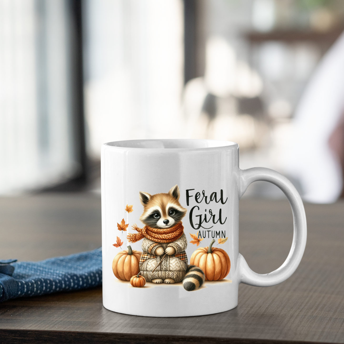 The Feral Girl Autumn Mug | Cozy Raccoon Fall Coffee Cup features a raccoon in a sweater and scarf with pumpkins and autumn leaves, adding a cozy autumn vibe to your morning coffee.