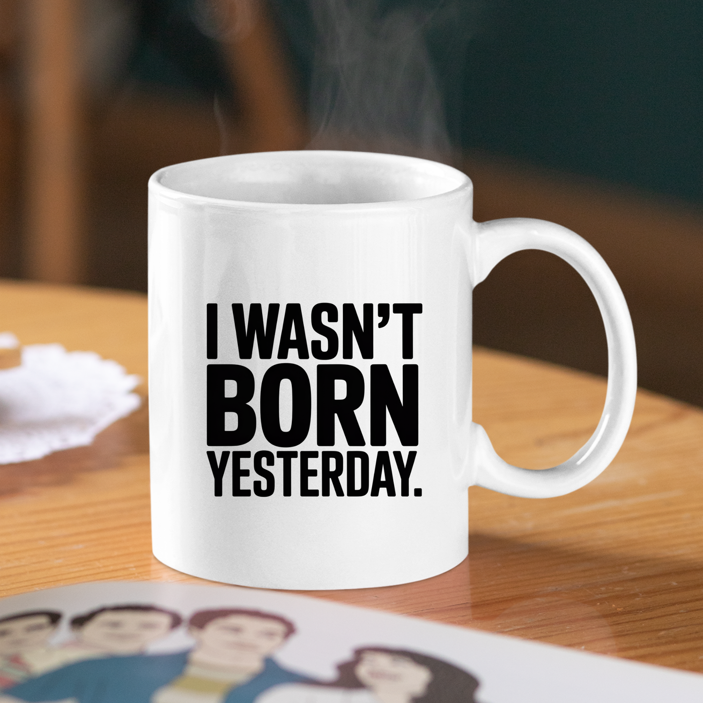 On a wooden table sits the "I Wasn't Born Yesterday Mug | Witty Quote Coffee Cup," a white ceramic mug featuring bold black text that reads "I WASN'T BORN YESTERDAY." Steam curls from the humorous coffee cup, while a partially visible photo or illustration rests nearby.