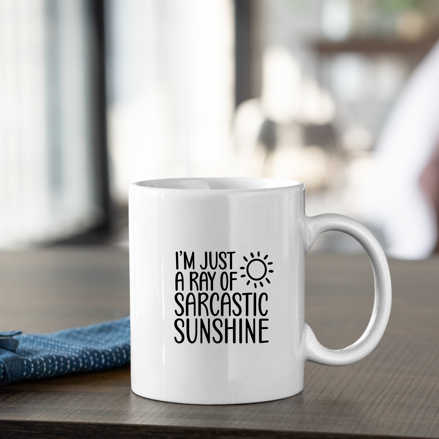 The Sarcastic Sunshine Mug | Funny Coffee Gift for Snarky People features “I’m just a ray of sarcastic sunshine” and a sun graphic—ideal for gifting coworkers and shown with a blue napkin in a softly lit room.