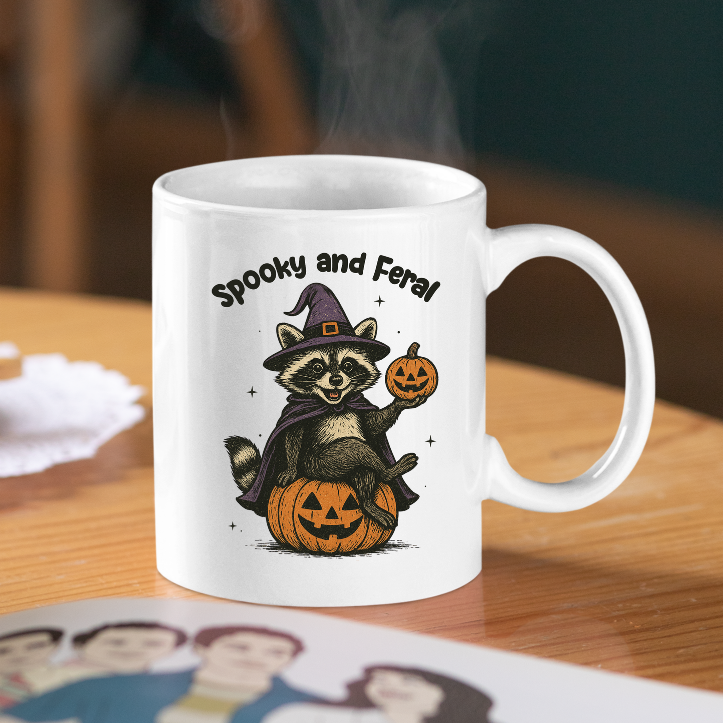 The Spooky and Feral Raccoon Mug | Funny Halloween Gift sits on a wooden table, showing a raccoon in a witch hat with pumpkins and the words "Spooky and Feral," steam rising from this fun Halloween mug.