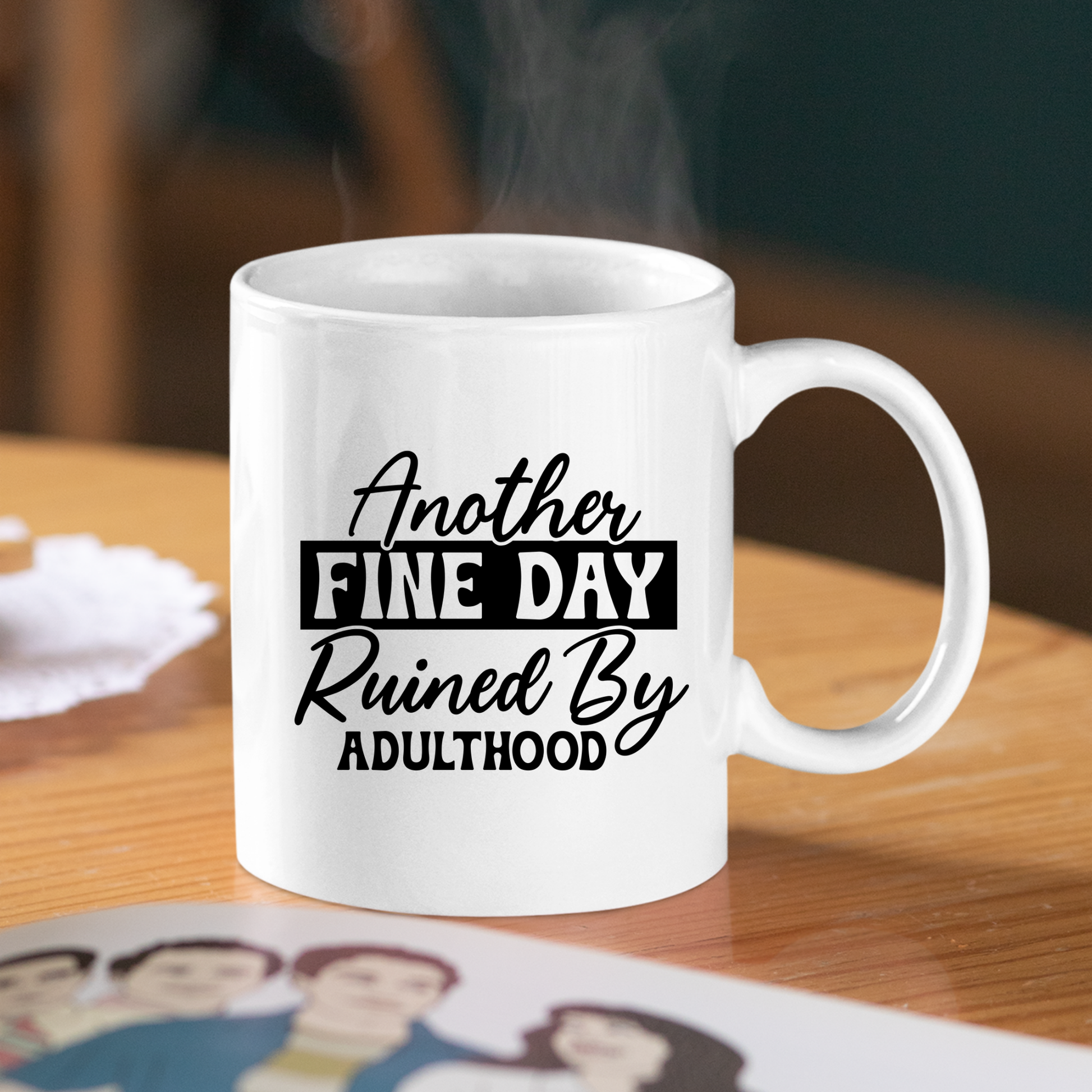 The Another Fine Day Mug – Funny Adulthood Coffee Cup sits on a wooden table, steam rising as its black text reads, "Another fine day ruined by adulthood." Ideal gift for new grads learning to adult; illustrated photo accents the scene.