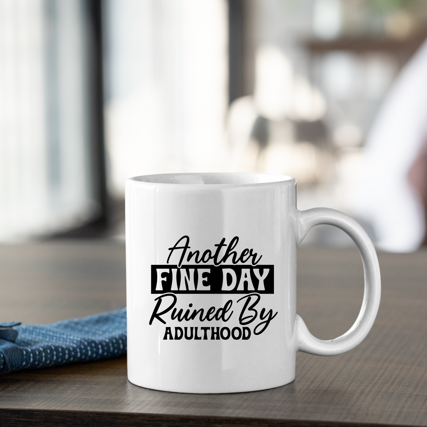 The Another Fine Day Mug – Funny Adulthood Coffee Cup features “Another fine day ruined by adulthood” in bold black letters, making it a perfect adulting mug or gift for new grads. It's pictured on a table with a blue cloth and blurred background.