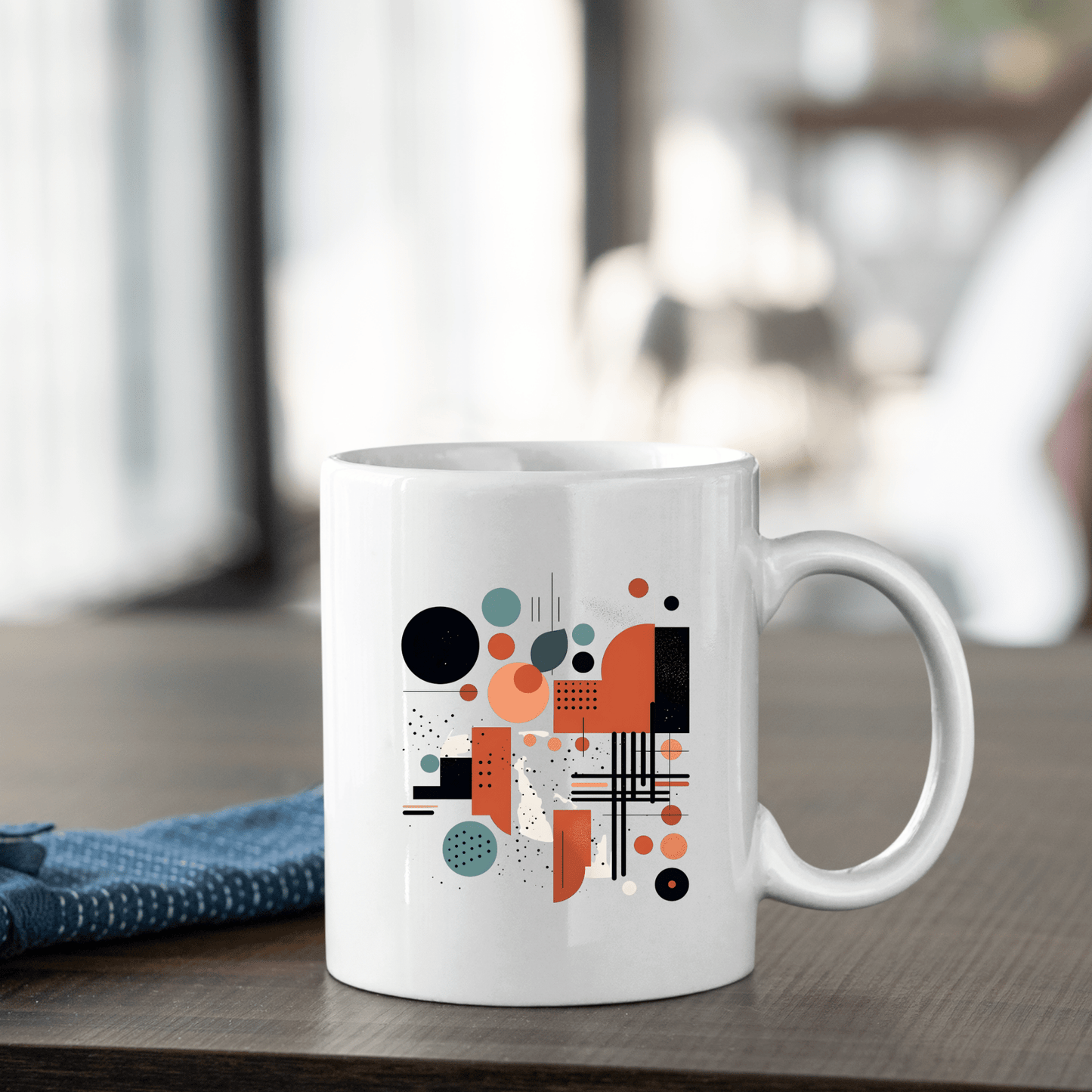 The Atomic Sunrise Mid Century Modern Mug, a white ceramic coffee cup with orange, black, and teal geometric circles and lines, rests on a wooden table beside a folded blue cloth. A softly blurred background enhances its stylish retro vibe.