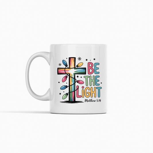 The Be the Light Matthew 5:14 Christmas Mug is a white coffee mug with a colorful cross wrapped in Christmas lights and "Be the Light Matthew 5:14" in playful, multicolored letters—perfect to inspire faith daily.