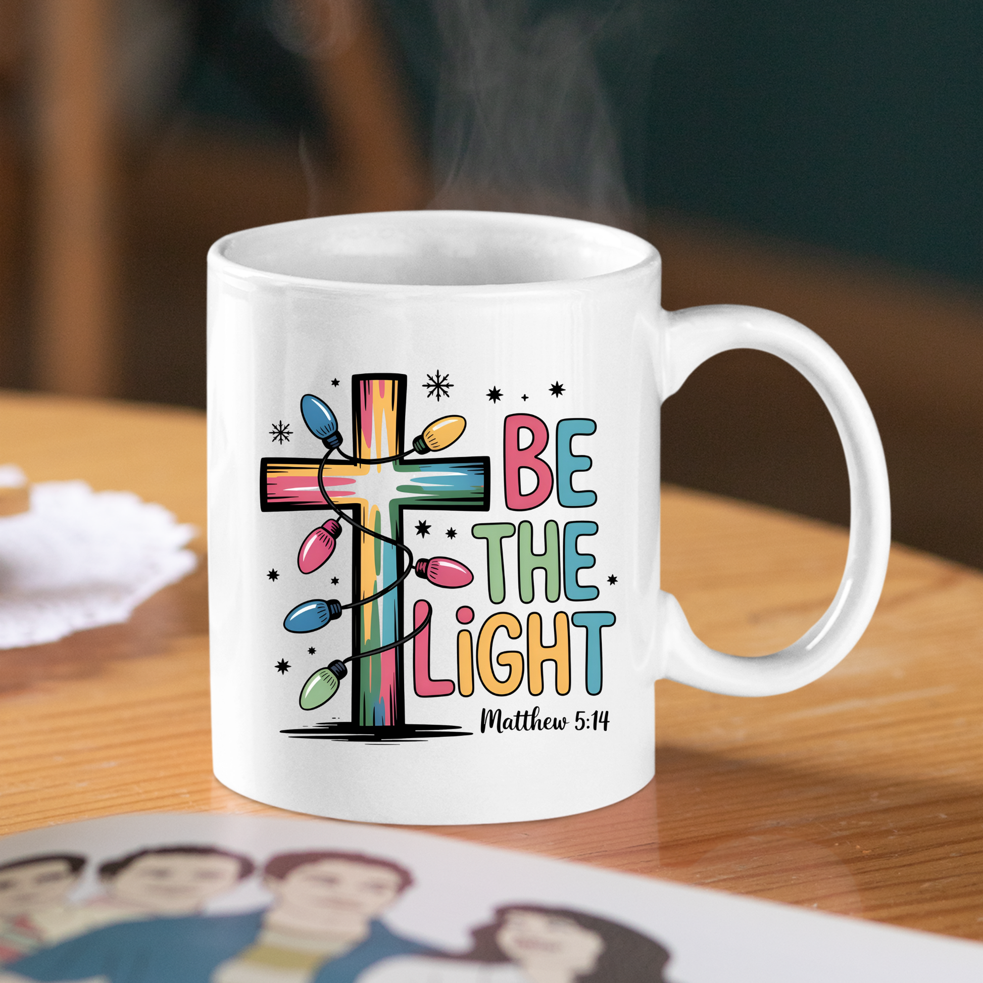 The Be the Light Matthew 5:14 Christmas Mug, adorned with a vibrant cross wrapped in Christmas lights and playful lettering, sits steaming on a wooden table—an ideal Christian faith gift for the season.