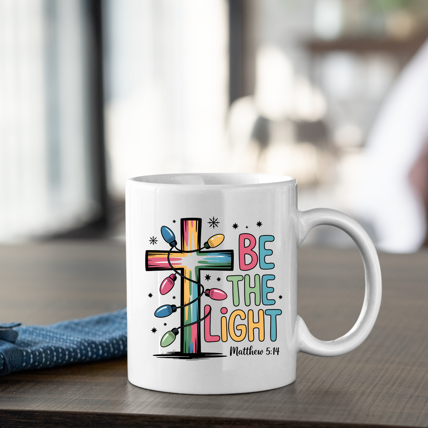 The Be the Light Matthew 5:14 Christmas Mug features a colorful cross wrapped in festive lights and “Be the Light, Matthew 5:14” lettering—the perfect faith-inspired gift for the holiday season.