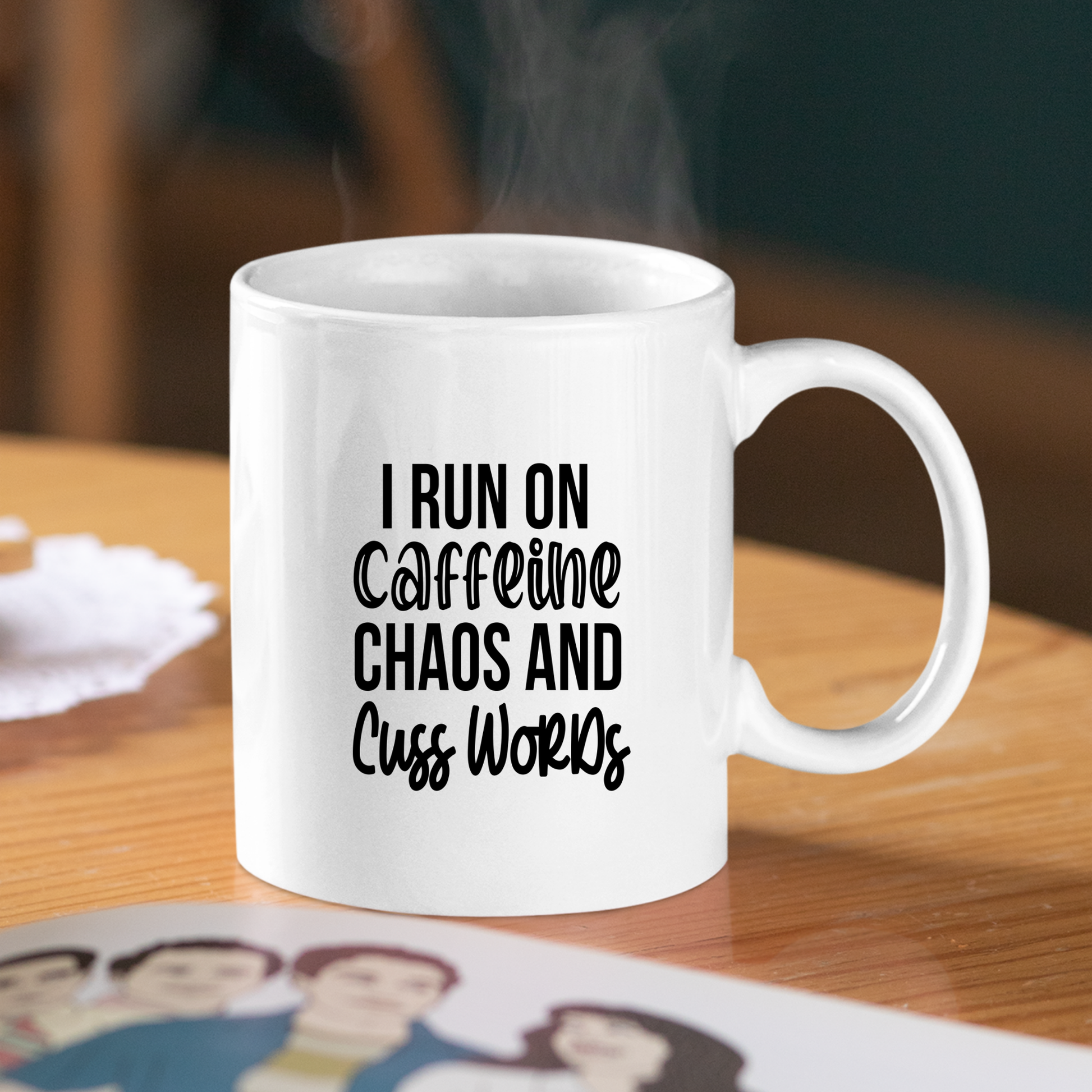 The Caffeine Chaos Cuss Words Mug | Funny Coffee Gift sits steaming on a wooden table with bold black text: “I RUN ON CAFFEINE, CHAOS AND CUSS WORDS,” while a nearby cartoon family photo adds a touch of charm to the scene.