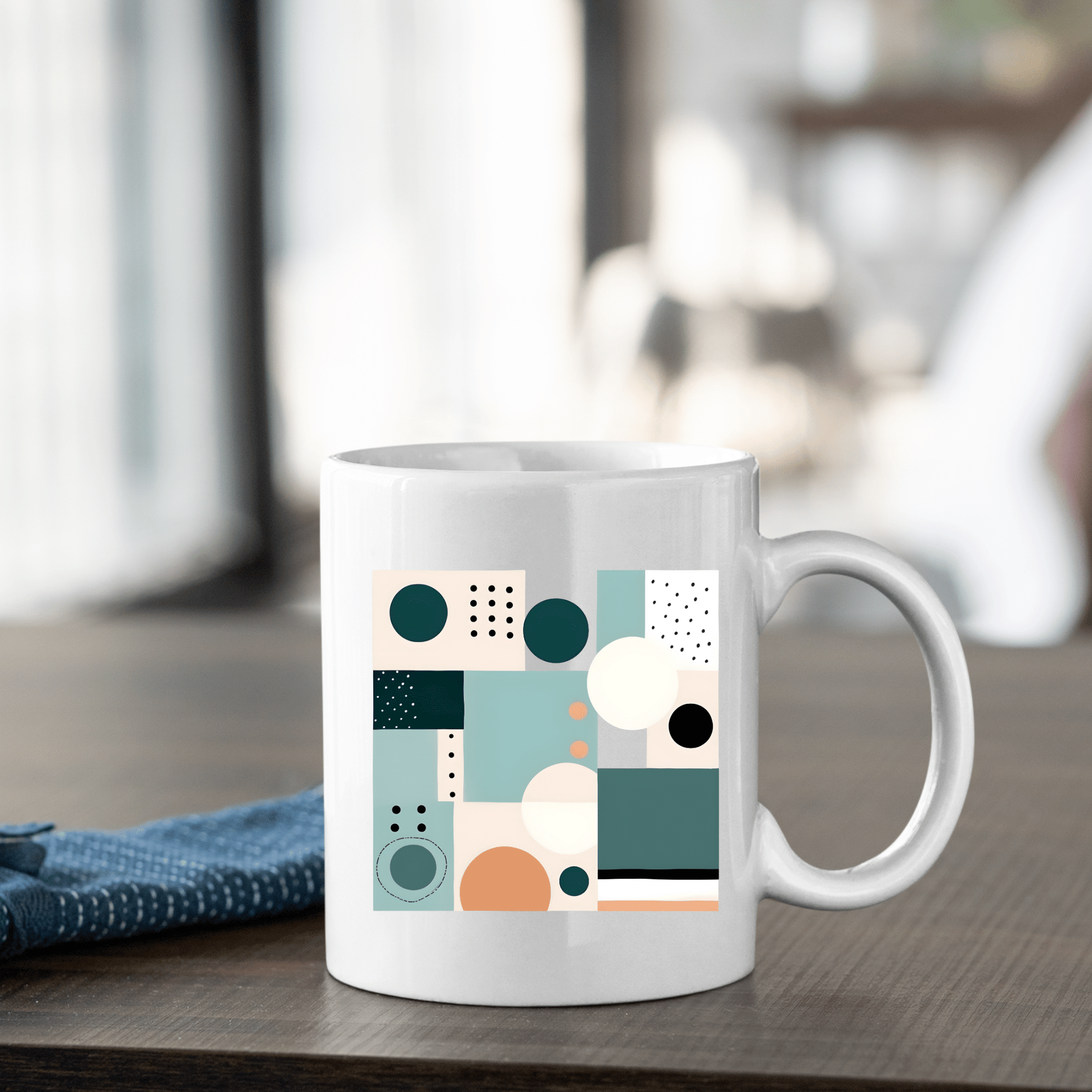 The Coastal Geometry Mid Century Modern Mug, a white ceramic mug with teal, peach, black, and white abstract shapes, sits on a wooden table beside a folded blue cloth.