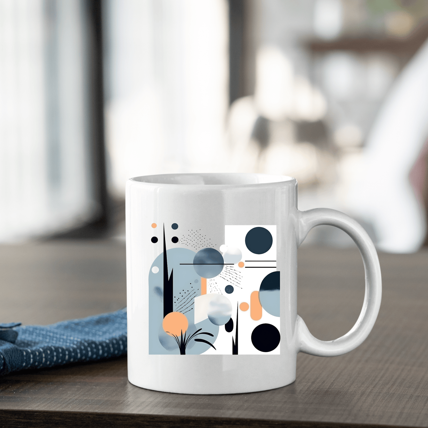 The Coastal Sunrise Mid Century Modern Mug, featuring a white ceramic body with abstract geometric designs in blue, black, orange, and gray, rests on a wooden table beside a folded blue cloth against a softly blurred background.