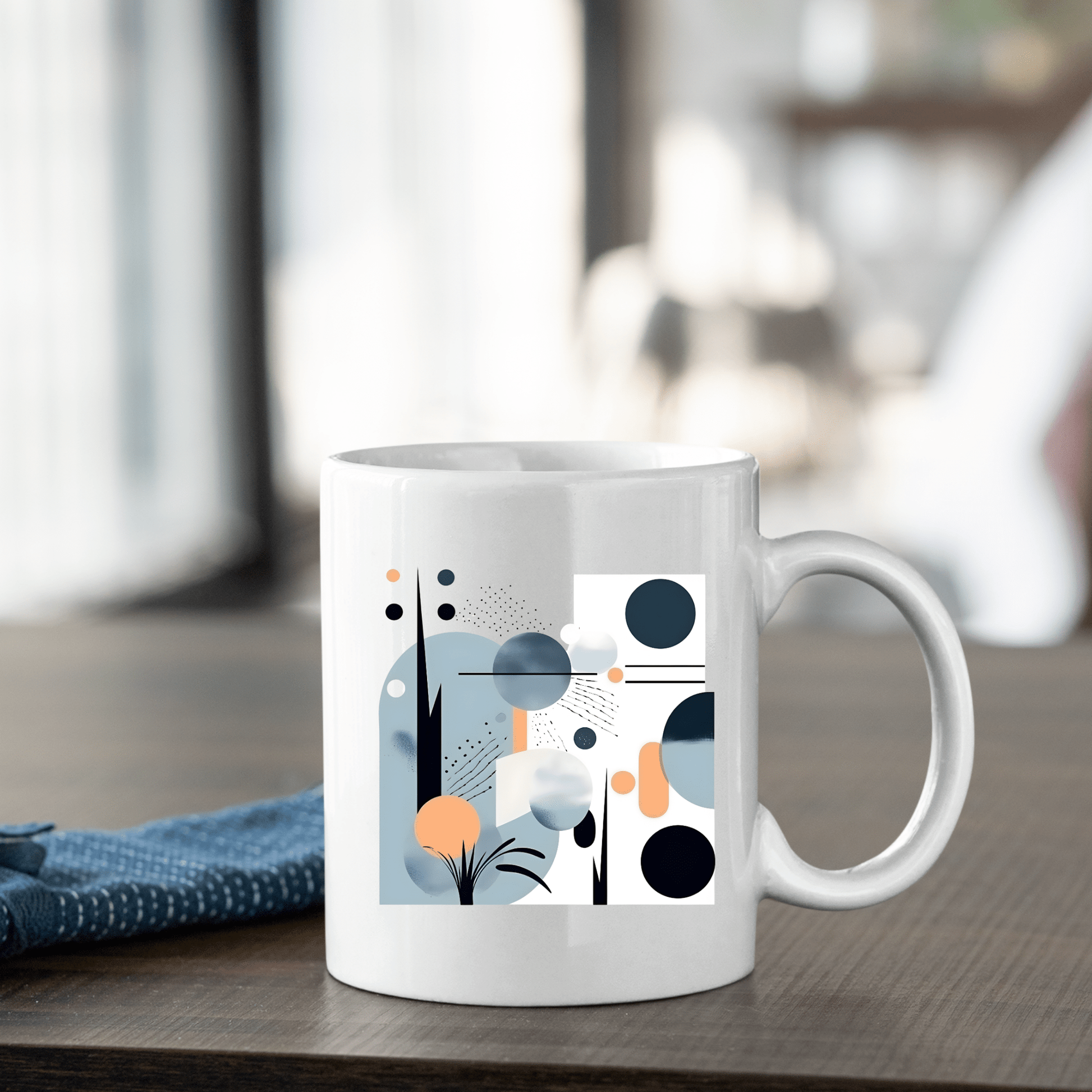The Coastal Sunrise Mid Century Modern Mug, featuring a white ceramic body with abstract geometric designs in blue, black, orange, and gray, rests on a wooden table beside a folded blue cloth against a softly blurred background.