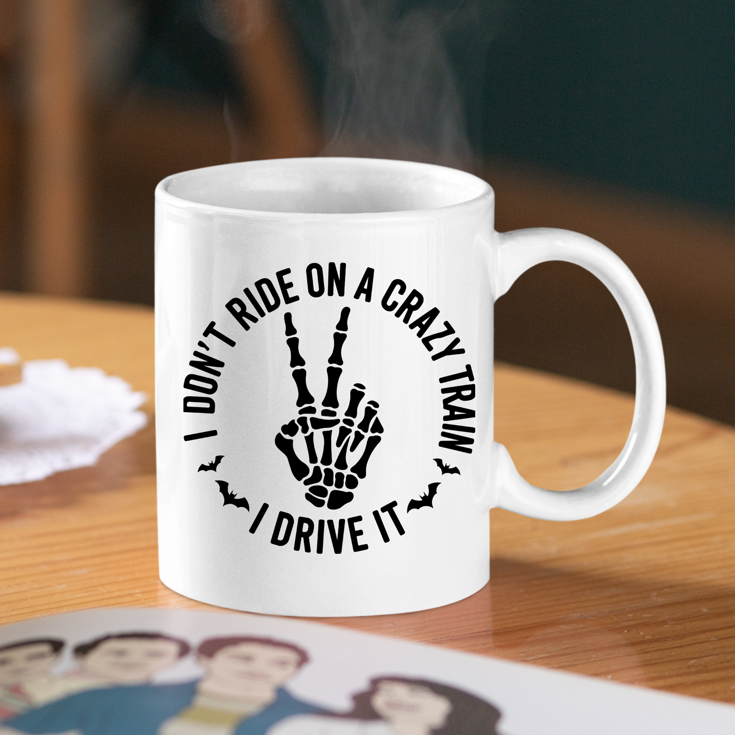 The Crazy Train Mug | Chaos Coffee Cup for Daily Mayhem features a skeleton hand flashing a peace sign with the phrase "I DON'T RIDE ON A CRAZY TRAIN, I DRIVE IT" encircled by playful bat illustrations. Resting on a wooden table, this darkly humorous mug is perfect for anyone ready to make a bold statement with their morning coffee.