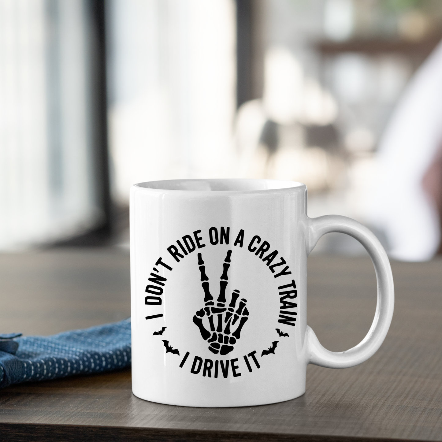 A Crazy Train Mug | Chaos Coffee Cup for Daily Mayhem sits on a wooden table, featuring a skeleton hand flashing the peace sign and the darkly humorous quote, “I don’t ride on a crazy train, I drive it,” surrounded by small bats. A blue cloth rests nearby.