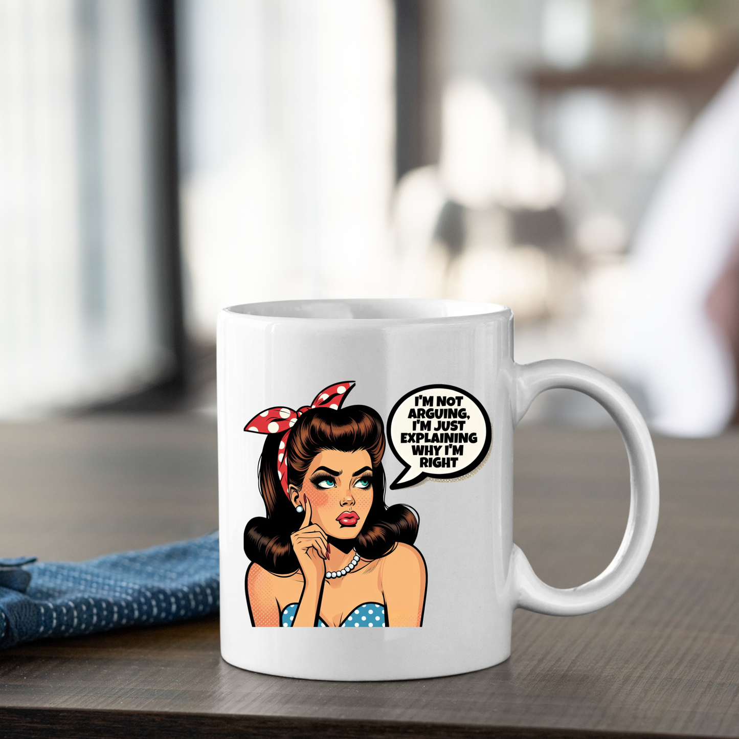 The Explaining Why I’m Right Mug | Funny Retro Coffee Gift sits on the table, featuring a vintage pop art woman with brown hair, a red polka-dot headband, and a speech bubble: “I’m not arguing, I’m just explaining why I’m right.”.