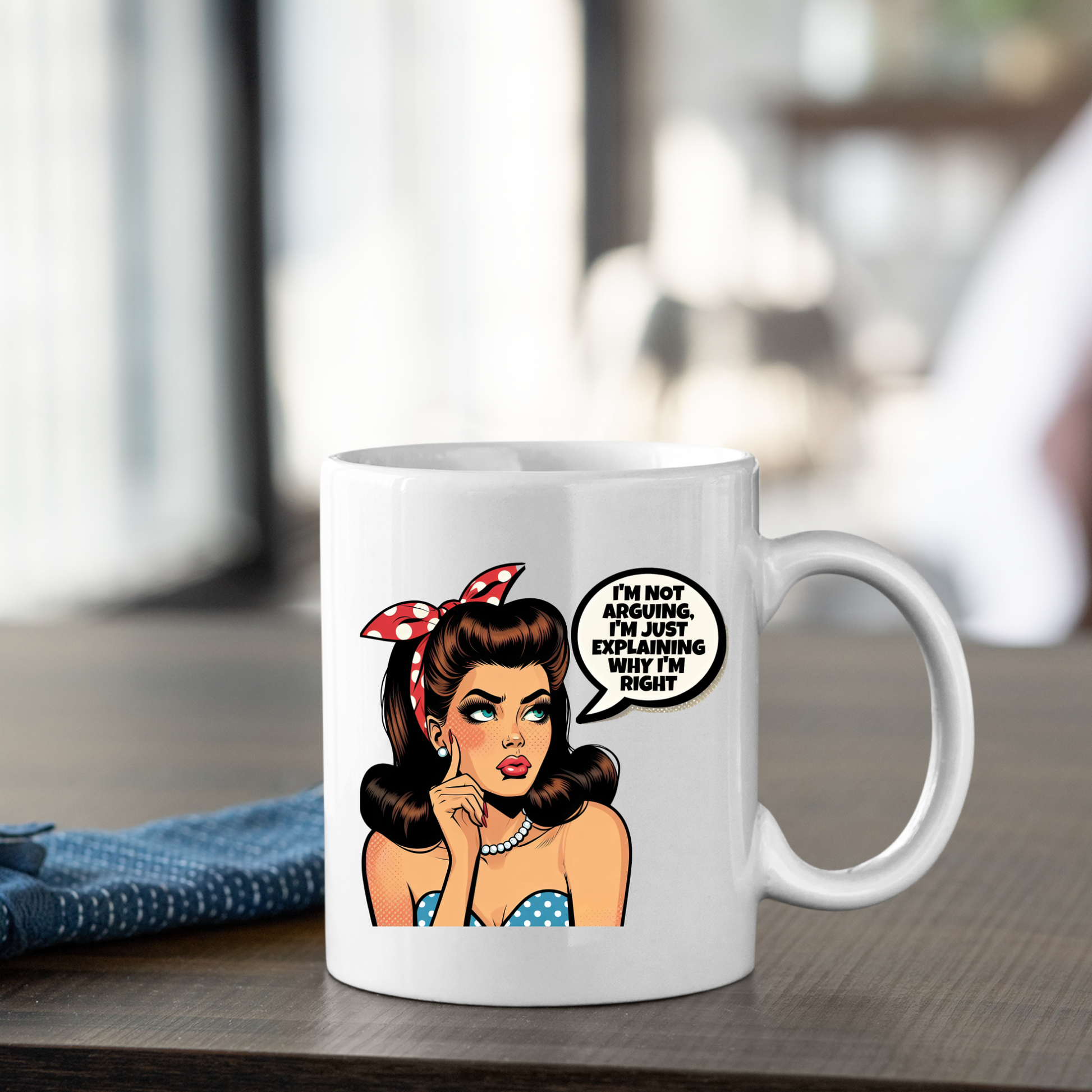 The Explaining Why I’m Right Mug | Funny Retro Coffee Gift sits on the table, featuring a vintage pop art woman with brown hair, a red polka-dot headband, and a speech bubble: “I’m not arguing, I’m just explaining why I’m right.”.