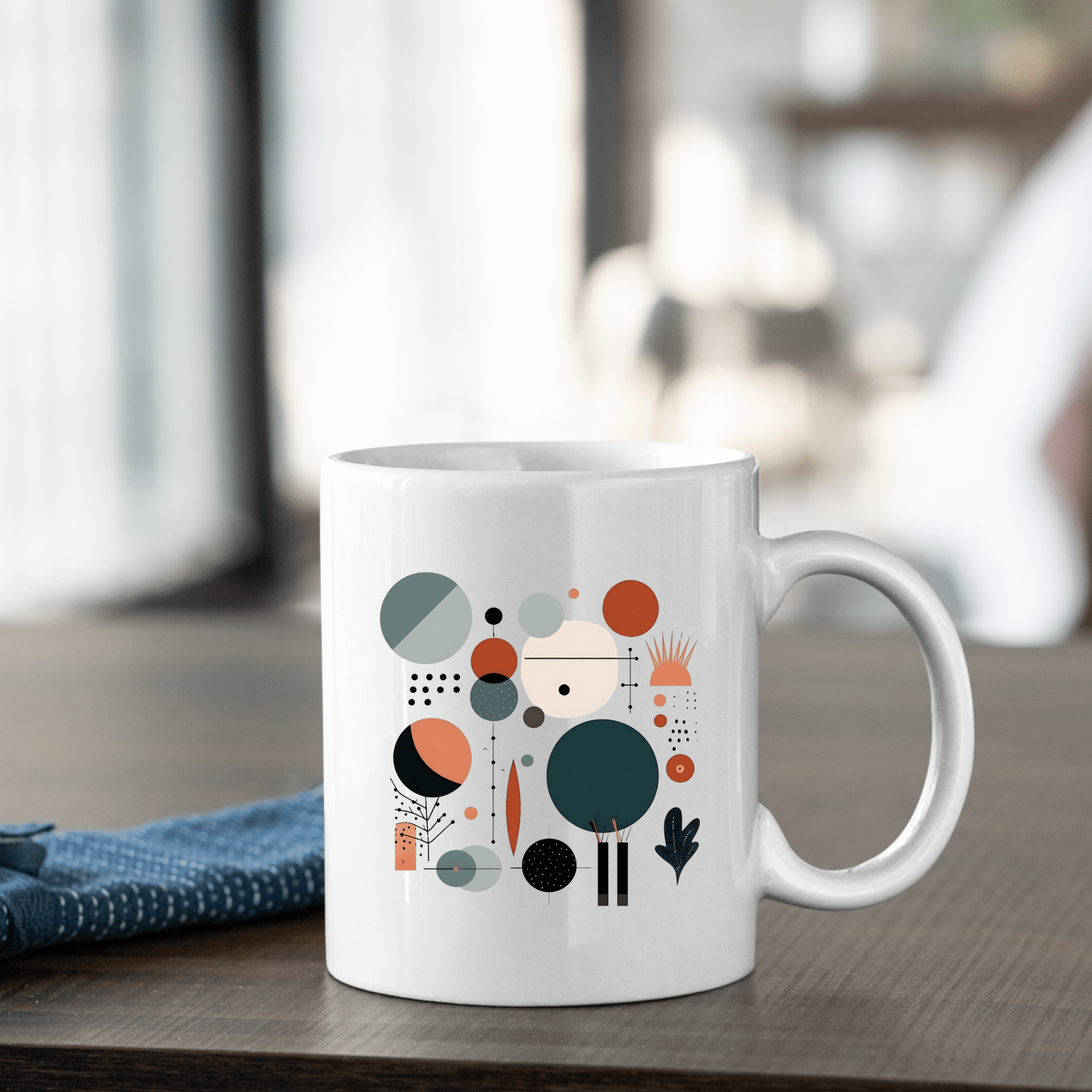 The Harvest Sun Mid Century Modern Mug, featuring a colorful abstract geometric design on white ceramic, sits on a wooden table beside a folded blue cloth with a blurred indoor background.
