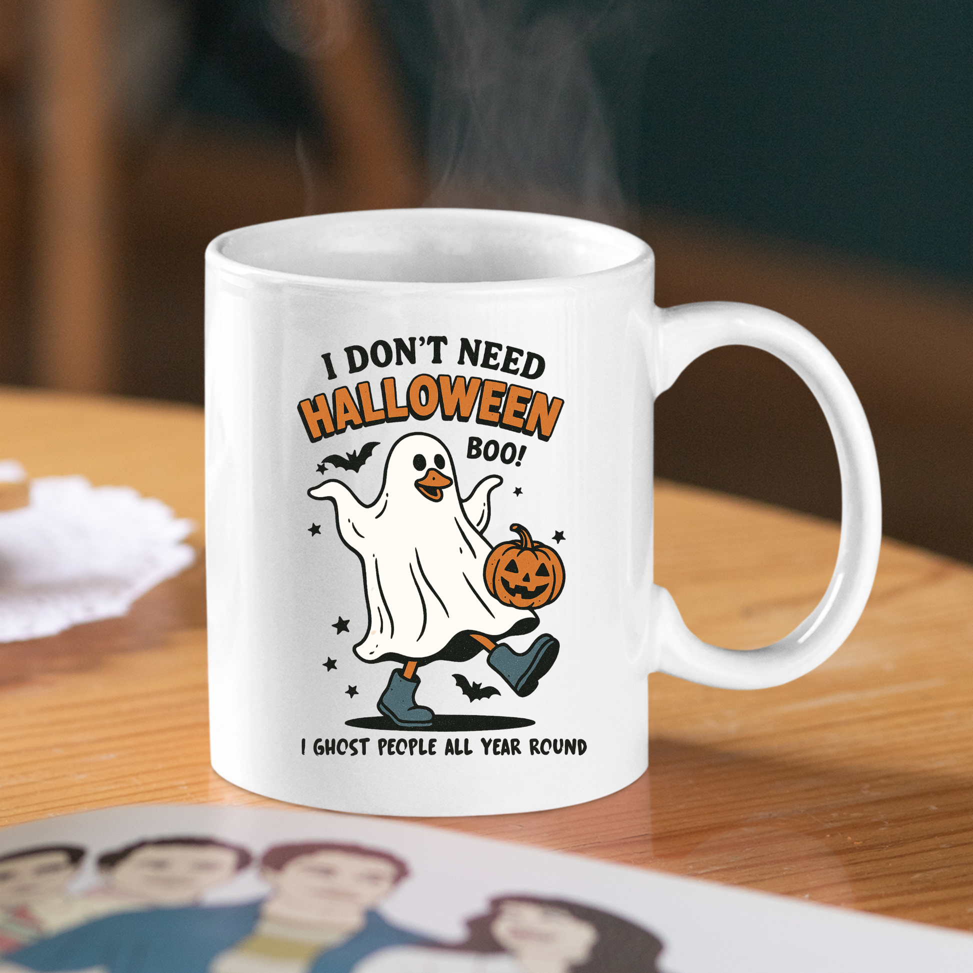 The Funny Ghost Mug | I Don’t Need Halloween Boo Cup sits on a wooden table, featuring a cartoon ghost with a jack-o'-lantern and bats. The mug reads: "I don't need Halloween boo! I ghost people all year round." Steam rises from the cup.