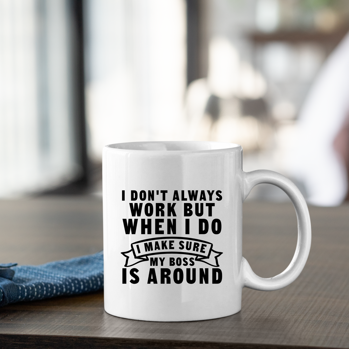The Funny Work Mug | I Make Sure My Boss Is Around sits on a wooden table, featuring its witty message in black text. A folded blue napkin is nearby, while the softly blurred background adds a charming touch.