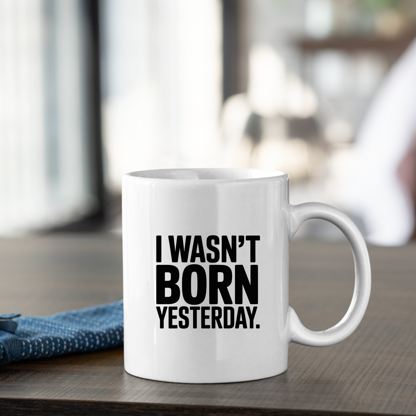 Certainly! Here’s the rewritten sentence using your product data: The I Wasn't Born Yesterday Mug | Witty Quote Coffee Cup is a white ceramic mug featuring "I WASN'T BORN YESTERDAY." printed in bold black letters. It sits on a wooden table beside a folded blue cloth, with the background softly blurred.