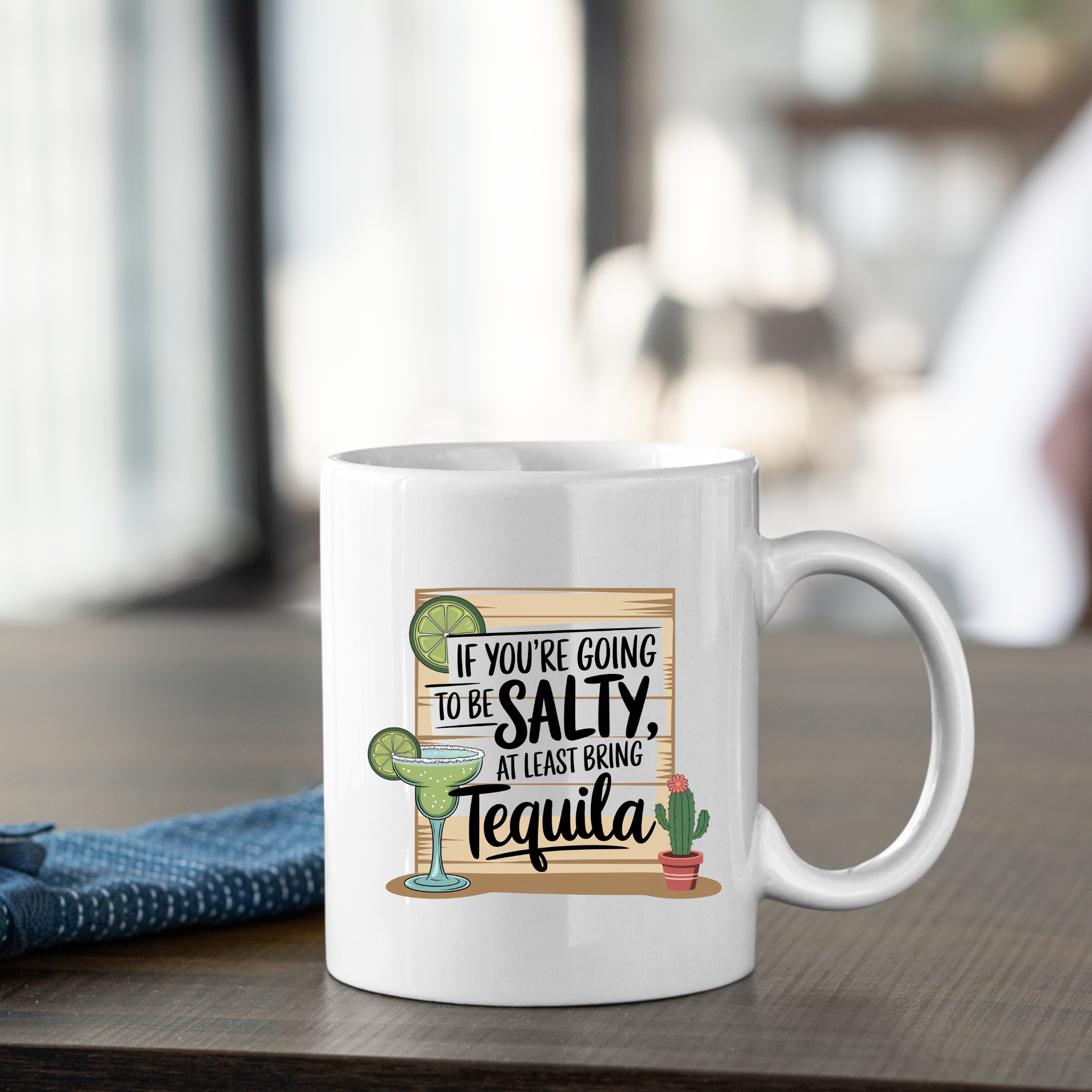 The If You're Going to Be Salty Bring Tequila Mug features a playful design with the phrase "If you're going to be salty, at least bring tequila," alongside illustrations of a margarita, lime, and cactus. This funny white mug makes the perfect tequila gift or margarita mug for anyone who loves a good laugh with their favorite drink.