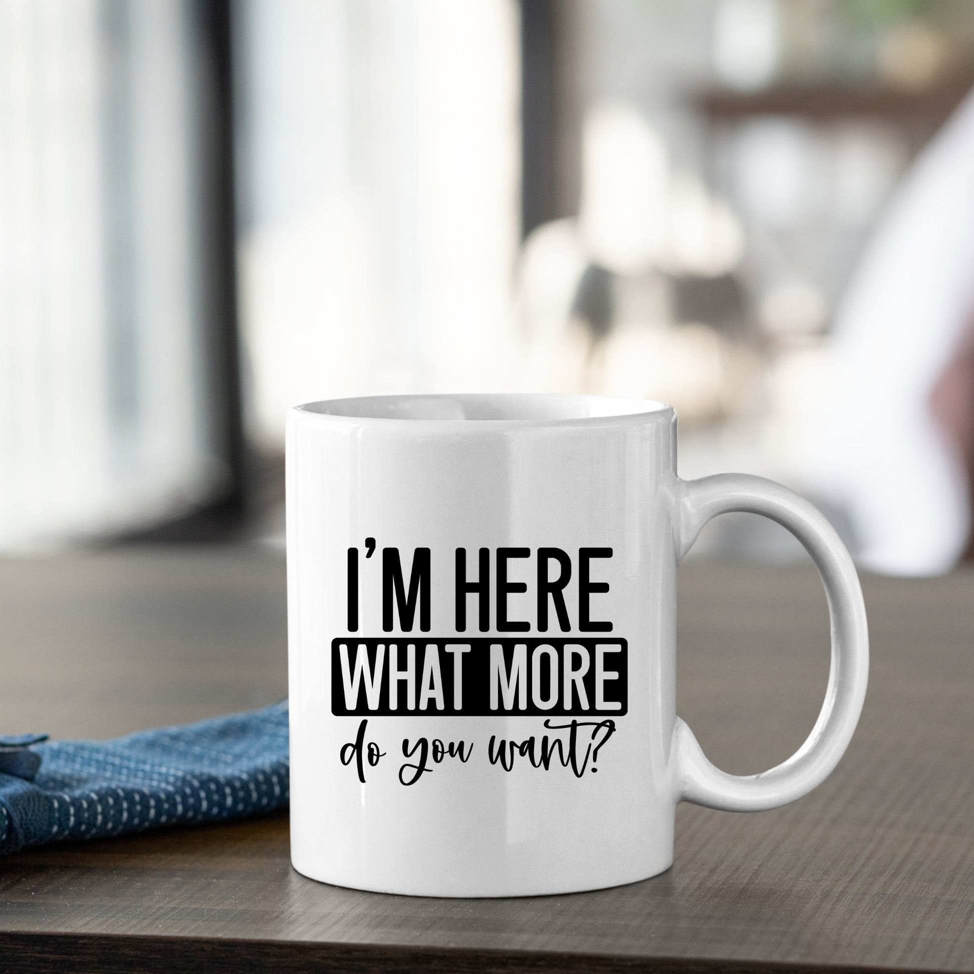 The “I’m Here What More Do You Want Mug | Funny Sarcastic Coffee Cup” sits on a wooden table with black text, great as an introvert gift or office mug, next to a folded blue cloth and soft, blurred background.