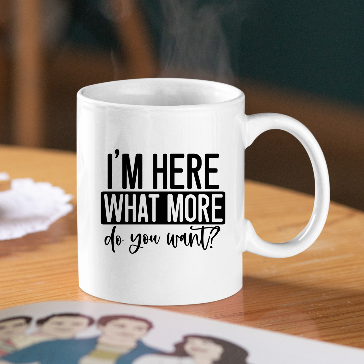 The "I’m Here What More Do You Want Mug" is a white, funny sarcastic coffee cup on a wooden table—steam rising, with a lace mat and photo nearby—making it the perfect introvert gift.