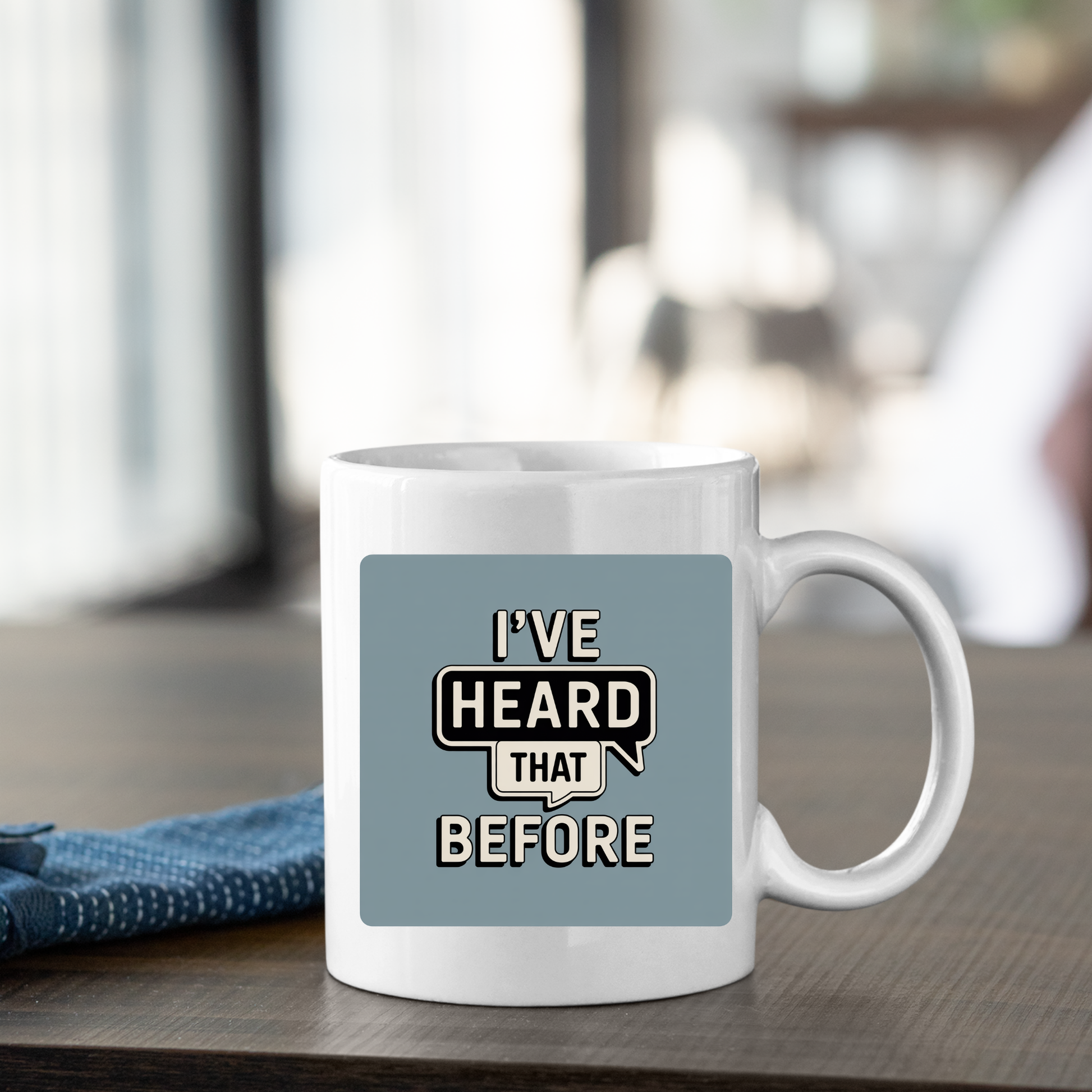 A white ceramic mug rests on a table beside a blue napkin. Featuring a bold label that reads "I'VE HEARD THAT BEFORE" on a gray background, the I've Heard That Before Mug | Office Humor Coffee Cup is the ideal choice for anyone who enjoys a touch of sarcasm and humor with their morning coffee.