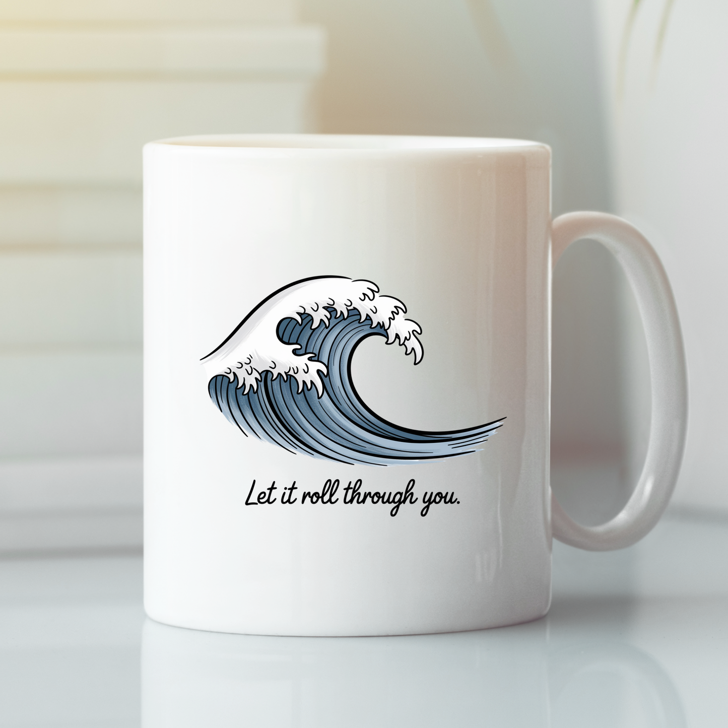 The "Let It Roll Through You Mug | Ocean Inspiration Gift" is a white coffee mug showcasing a blue ocean wave illustration with the phrase "Let it roll through you." printed beneath the wave, displayed on a white surface against a softly blurred background.