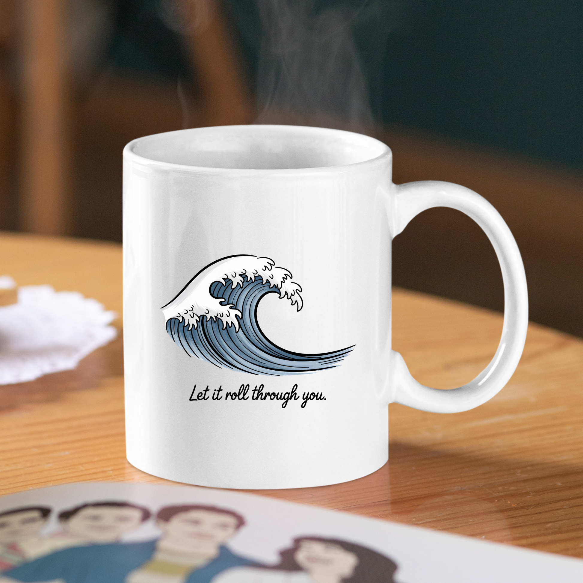 The Let It Roll Through You Mug | Ocean Inspiration Gift, adorned with a blue wave illustration and the phrase "Let it roll through you," sits on a wooden table beside a blurred photo of people and a lace coaster. Steam rises gently from this inspirational coastal ceramic mug, adding warmth to the tranquil scene.