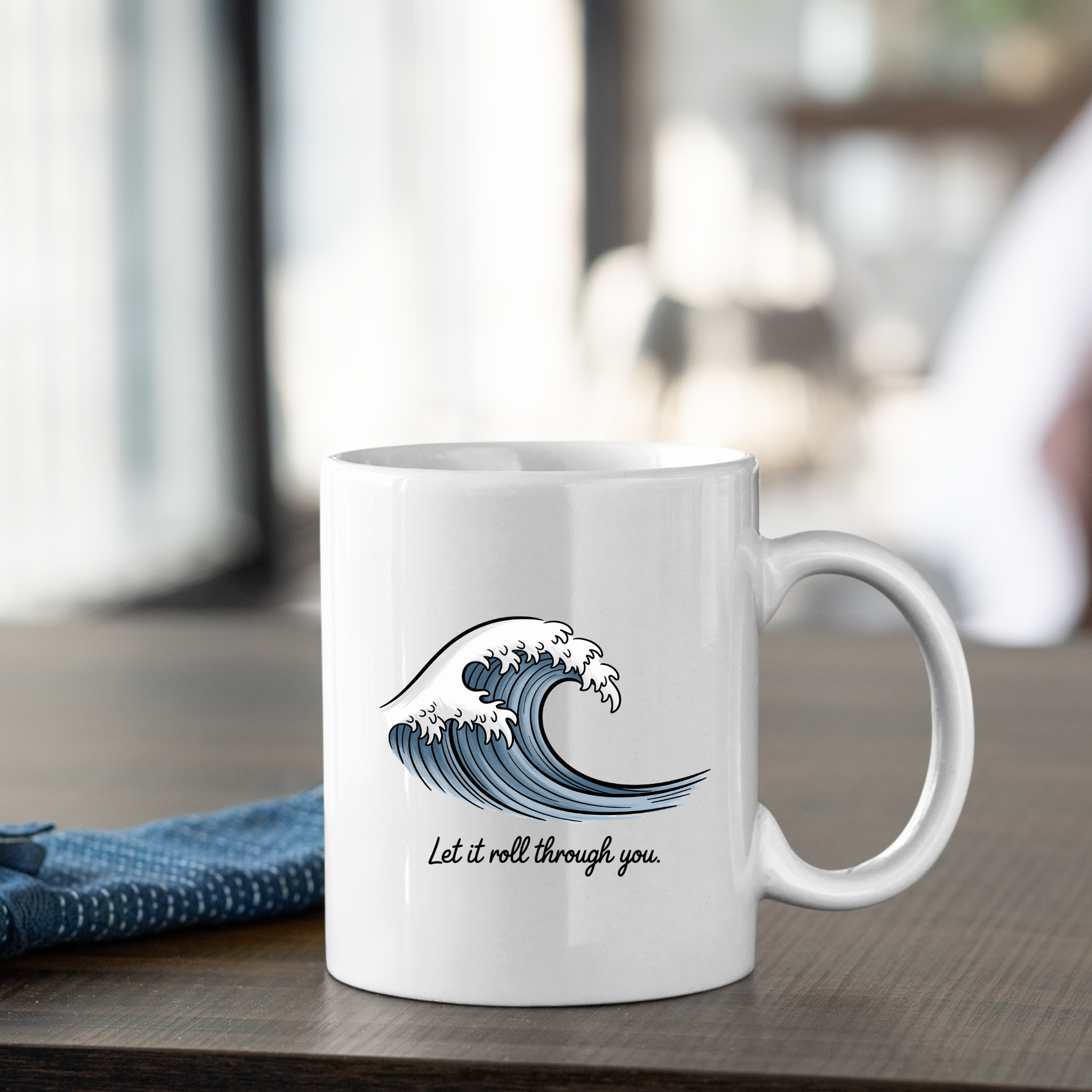 The Let It Roll Through You Mug | Ocean Inspiration Gift, a white ceramic mug adorned with a blue wave illustration and the phrase "Let it roll through you," rests on a wooden table next to a folded blue cloth, set against a softly blurred indoor background.