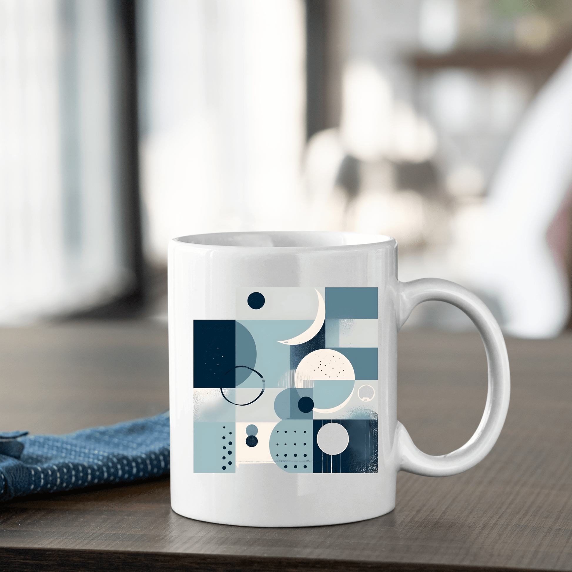 The Moonlit Geometry Mid Century Modern Mug, featuring an abstract geometric design in blue and gray, rests on a wooden table beside a folded blue cloth, set against a softly blurred background.