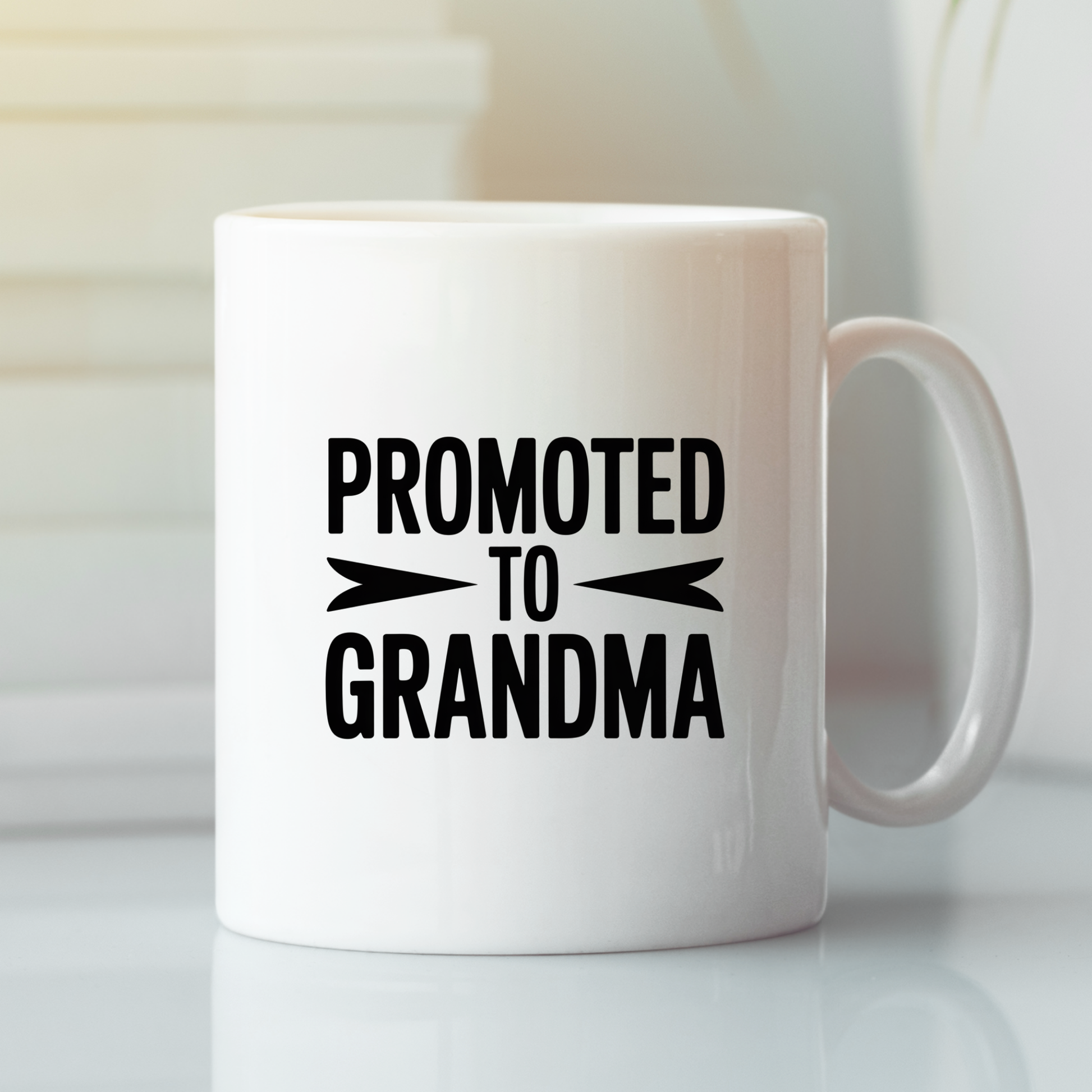 A Promoted to Grandma Mug | New Grandma Gift featuring bold black text that reads "PROMOTED TO GRANDMA" rests on a gleaming white surface against a softly blurred background. This charming mug is an ideal way to announce a pregnancy or surprise a loved one with the perfect grandma gift for your exciting news.