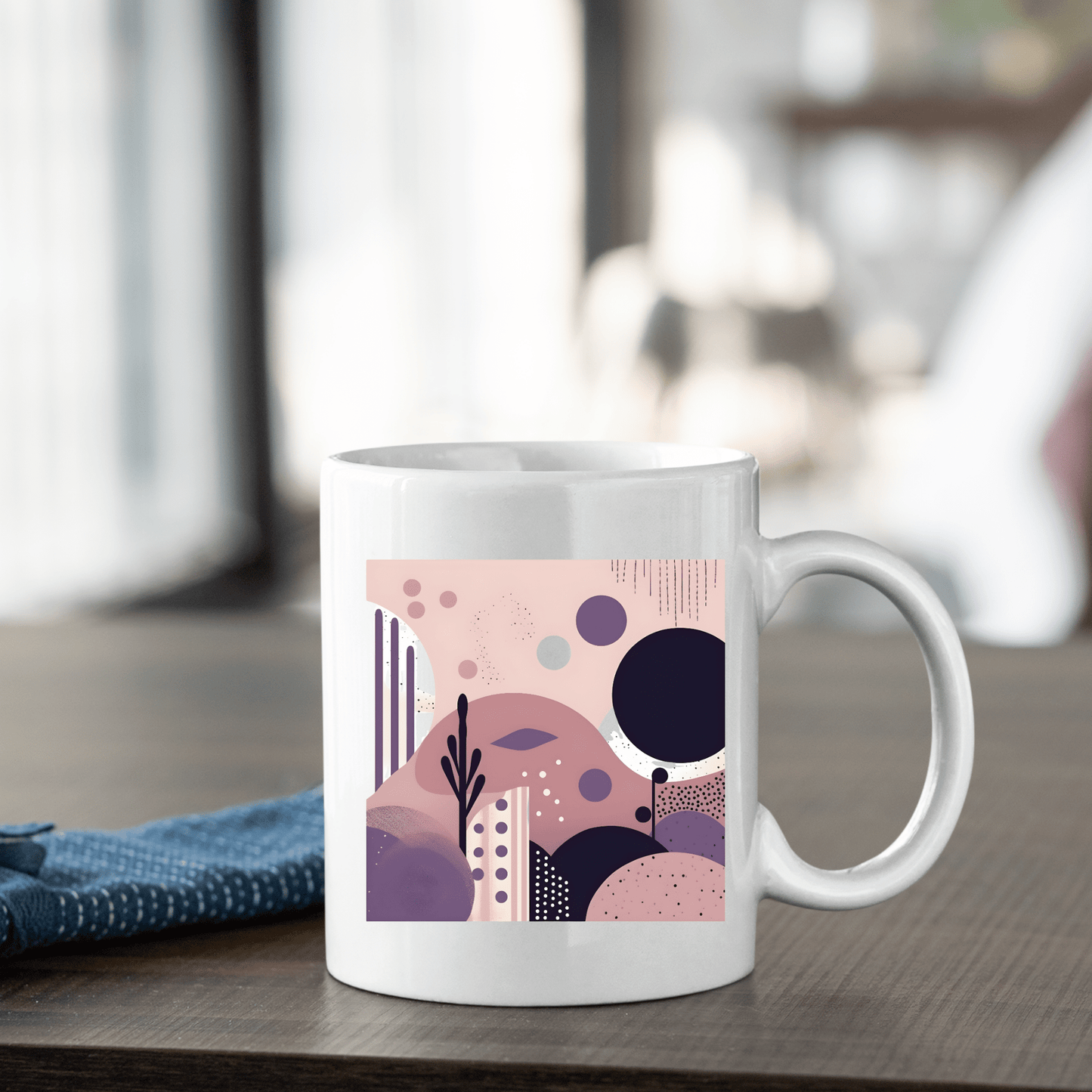 The Purple Dusk Mid Century Modern Mug, a white ceramic mug with an abstract pink, purple, and blue design, rests on a wooden table beside a folded dark blue cloth against a softly blurred bright background.