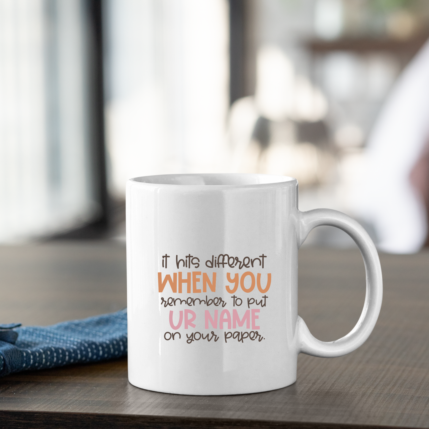 The Put Your Name On Your Paper Mug | Funny Teacher Gift sits on a wooden table with the text: “It hits different WHEN YOU remember to put UR NAME on your paper,” a great gift for teachers, blue napkin beside it and a softly blurred background.