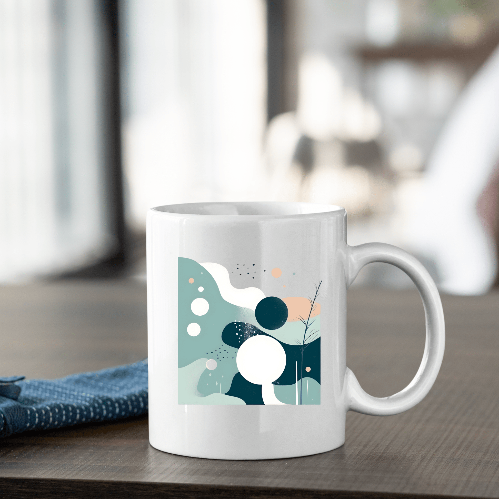 The Quiet Tides Mid Century Modern Mug, featuring a white ceramic body with abstract geometric designs in blue, green, and peach tones, sits on a wooden table beside a folded blue cloth—a stylish piece of minimalist home decor.