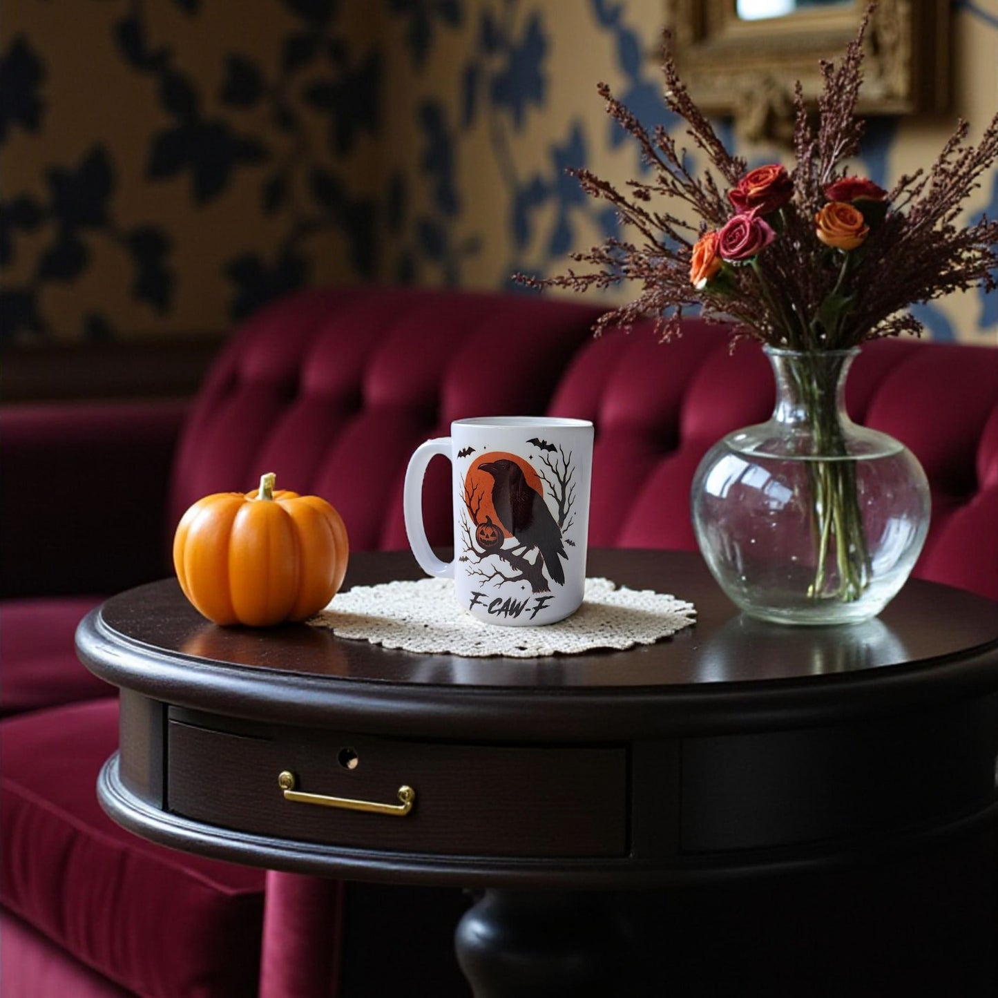 The F-CAW-F Raven Mug | Gothic Halloween Coffee Cup sits on a dark table with an orange pumpkin and dried flowers, creating a spooky fall vibe—perfect as a Halloween gift or autumn decor accessory.