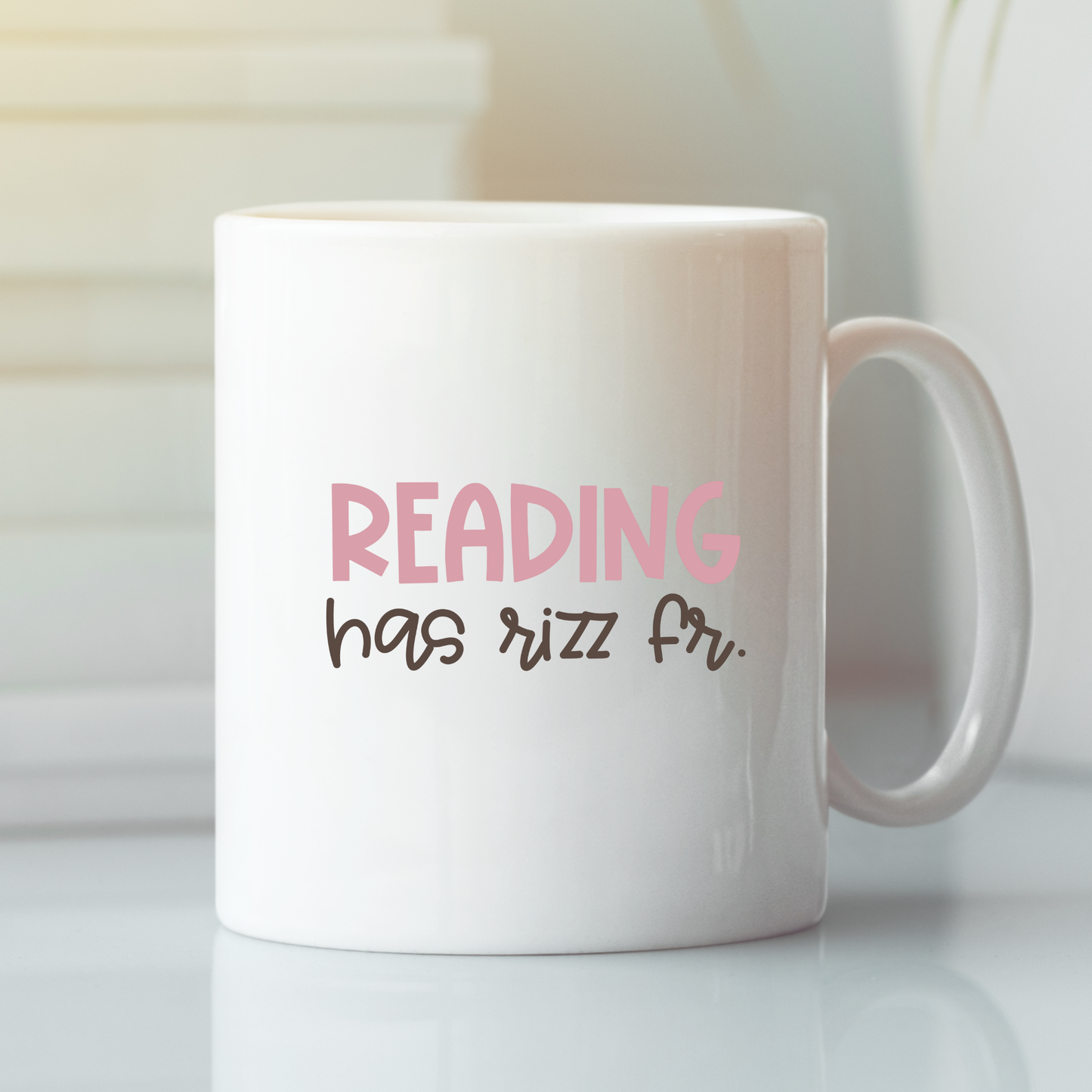 The Reading Has Rizz Mug | Funny Literacy Teacher Gift features "READING has rizz fr." in pink and black on white—ideal for teacher appreciation or book lovers, pictured on a light surface with books blurred in the background.