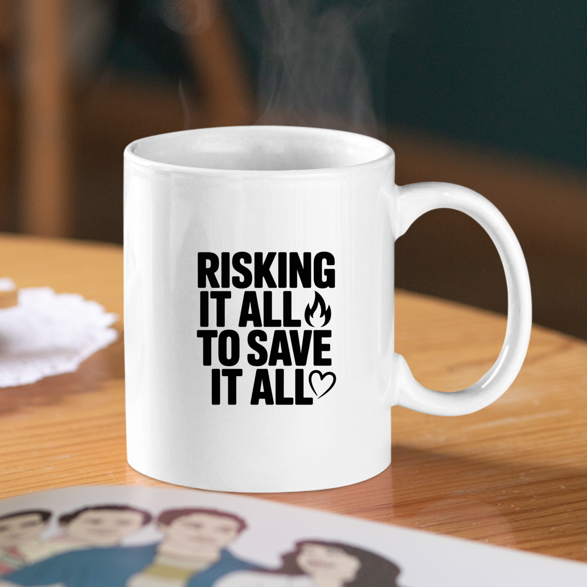 The Risking It All to Save It All Firefighter Mug—a white ceramic mug featuring the bold black phrase "RISKING IT ALL TO SAVE IT ALL," highlighted by a small flame icon and heart symbol—rests on a wooden table beside a coaster and a photo. Steam curls upwards, making it the perfect firefighter gift and tribute to courage.