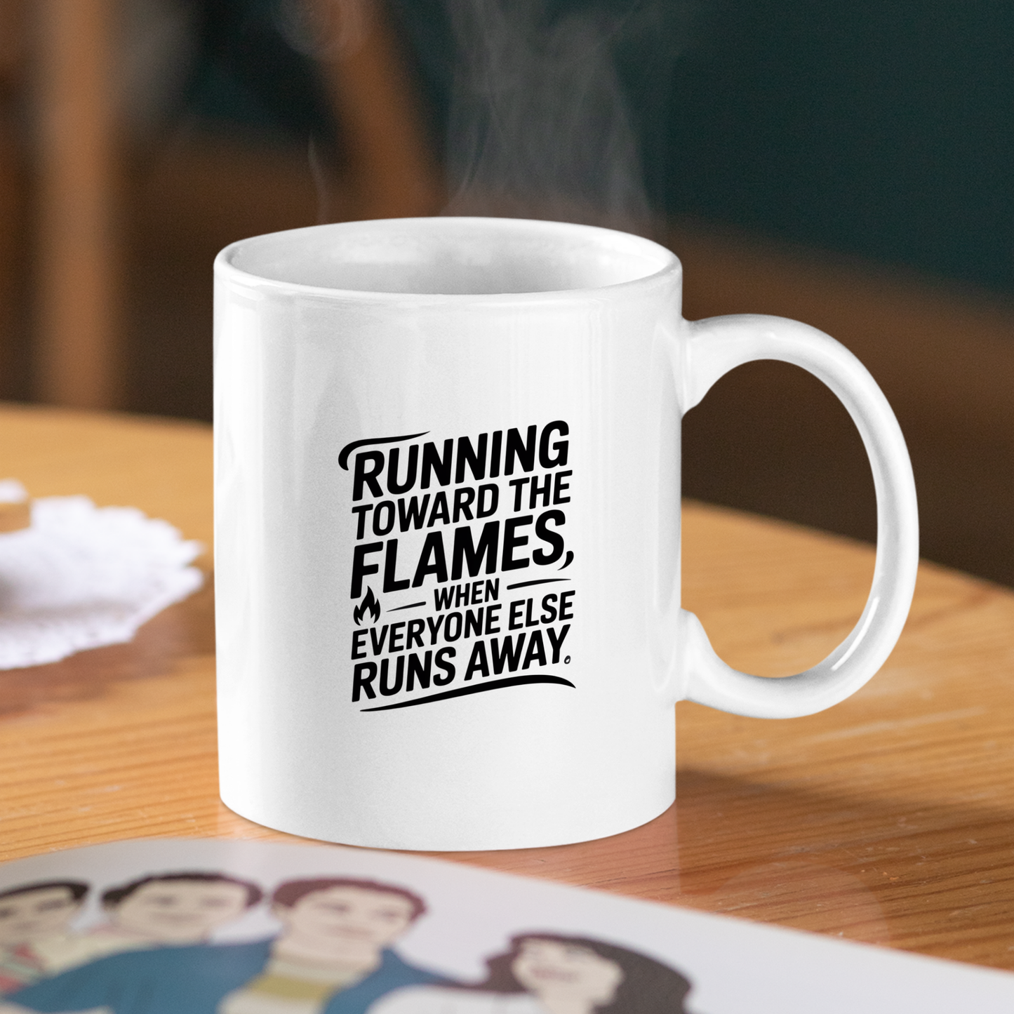 A Running Toward the Flames Firefighter Mug sits on a table with steam rising from it. This white mug features bold black text: “Running toward the flames, when everyone else runs away.” Beside the mug—a perfect firefighter gift—a blurred photo or card is partially visible.