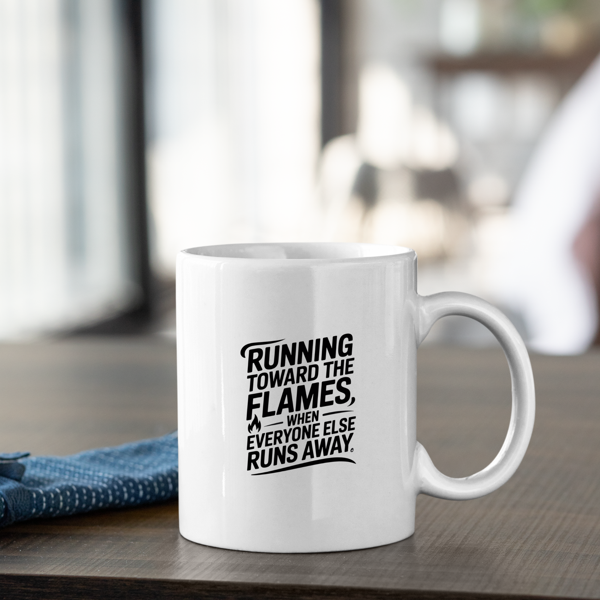 The Running Toward the Flames Firefighter Mug, in white, rests on a wooden table featuring the inspiring text: “Running toward the flames, when everyone else runs away.” A neatly folded blue cloth lies beside it, with a softly blurred background—making this courage-themed mug an ideal firefighter gift.