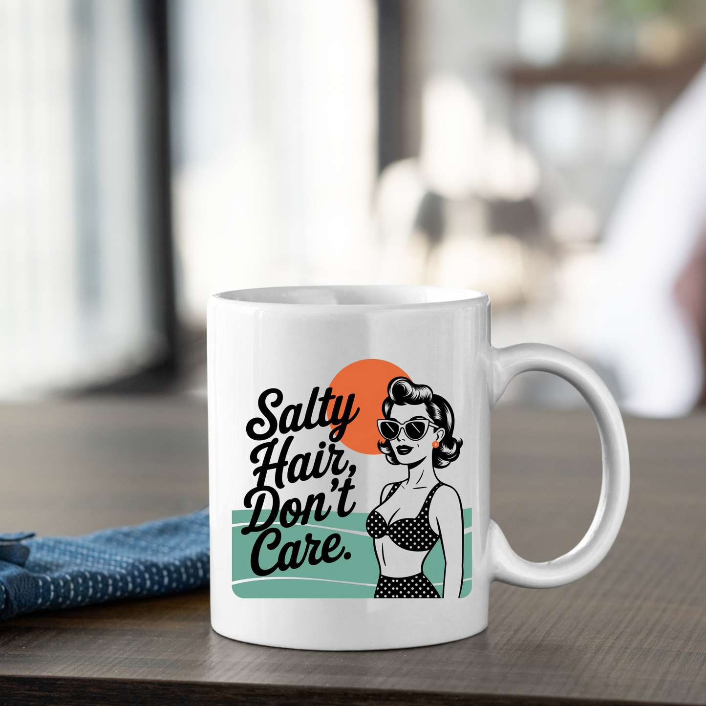 Enjoy your coffee in the Salty Hair Don't Care Mug | Retro Beach Coffee Mug, featuring a vintage-inspired illustration of a woman in sunglasses and a polka dot bikini with the playful phrase "Salty Hair, Don't Care." Ideal as a beach mug or coastal gift, this stylish mug is pictured sitting on a wooden table next to a blue cloth.