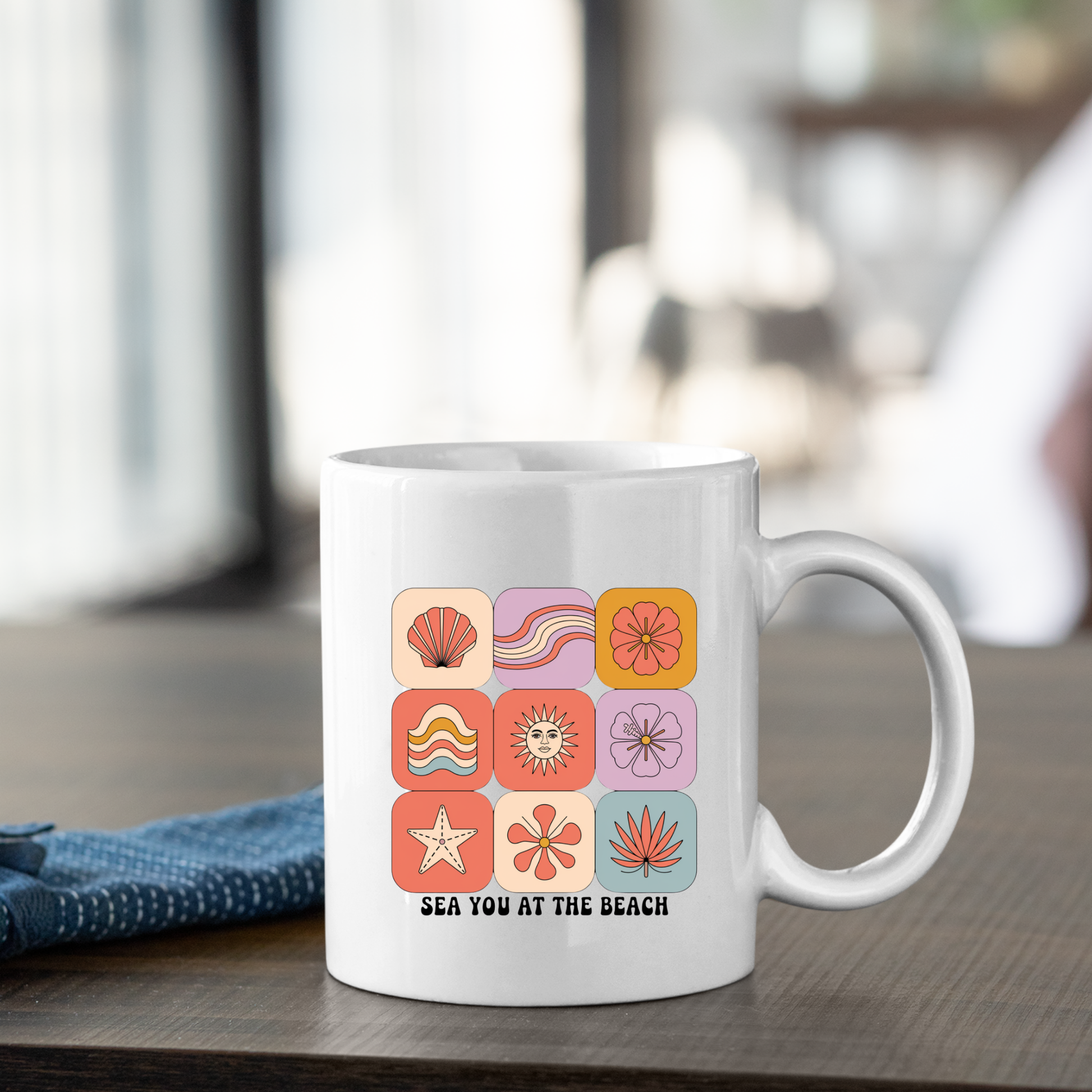 The Sea You at the Beach Mug | Retro Beach Coffee Mug, featuring colorful beach-themed icons and the phrase "SEA YOU AT THE BEACH," sits on a wooden table next to a folded blue napkin.