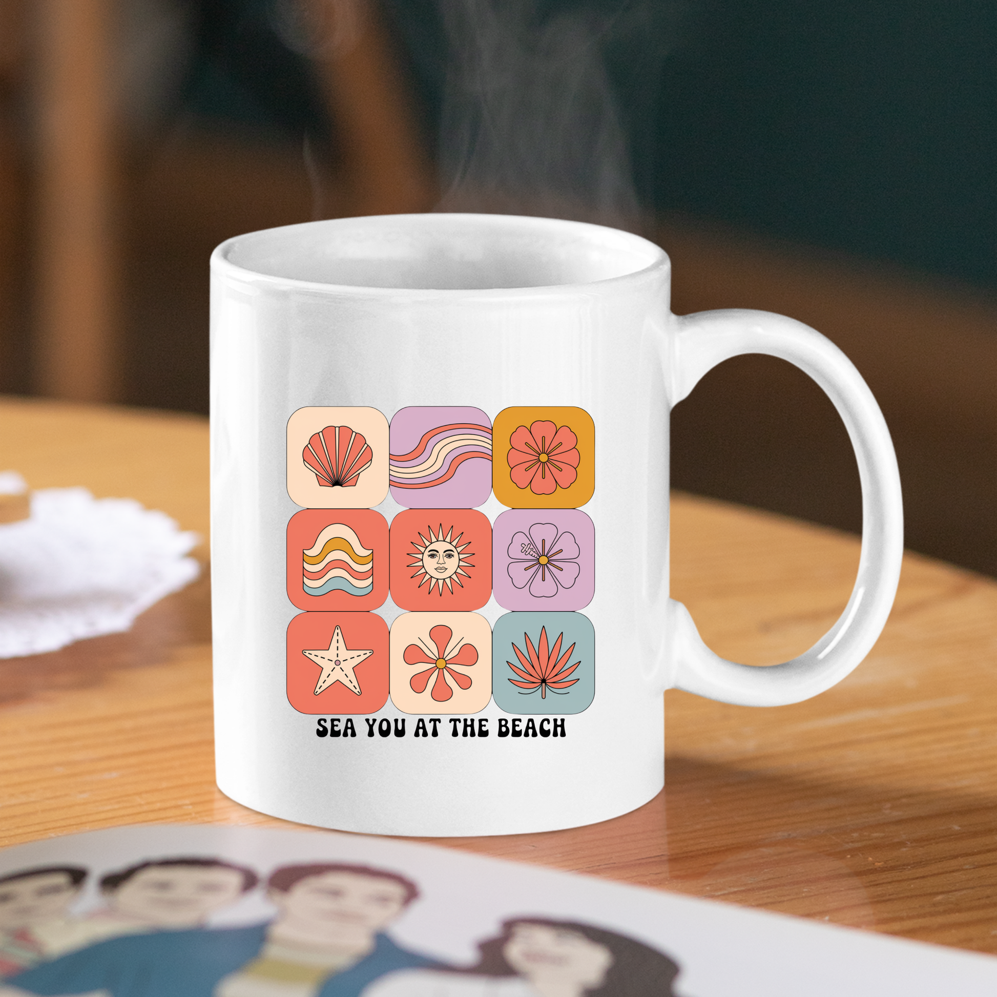The Sea You at the Beach Mug | Retro Beach Coffee Mug features a white coastal design adorned with colorful beach-themed icons—shells, waves, sun, and starfish—and cheerful text that reads "SEA YOU AT THE BEACH." Perfect for summer mornings, this charming mug sits invitingly on a wooden table next to your favorite book or magazine as steam gently rises from your beverage.