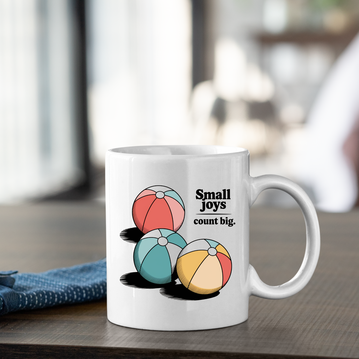 The Small Joys Count Big Mug | Inspirational Coffee Mug, featuring three colorful beach balls and the phrase "Small joys count big," sits on a wooden table near a folded blue napkin.