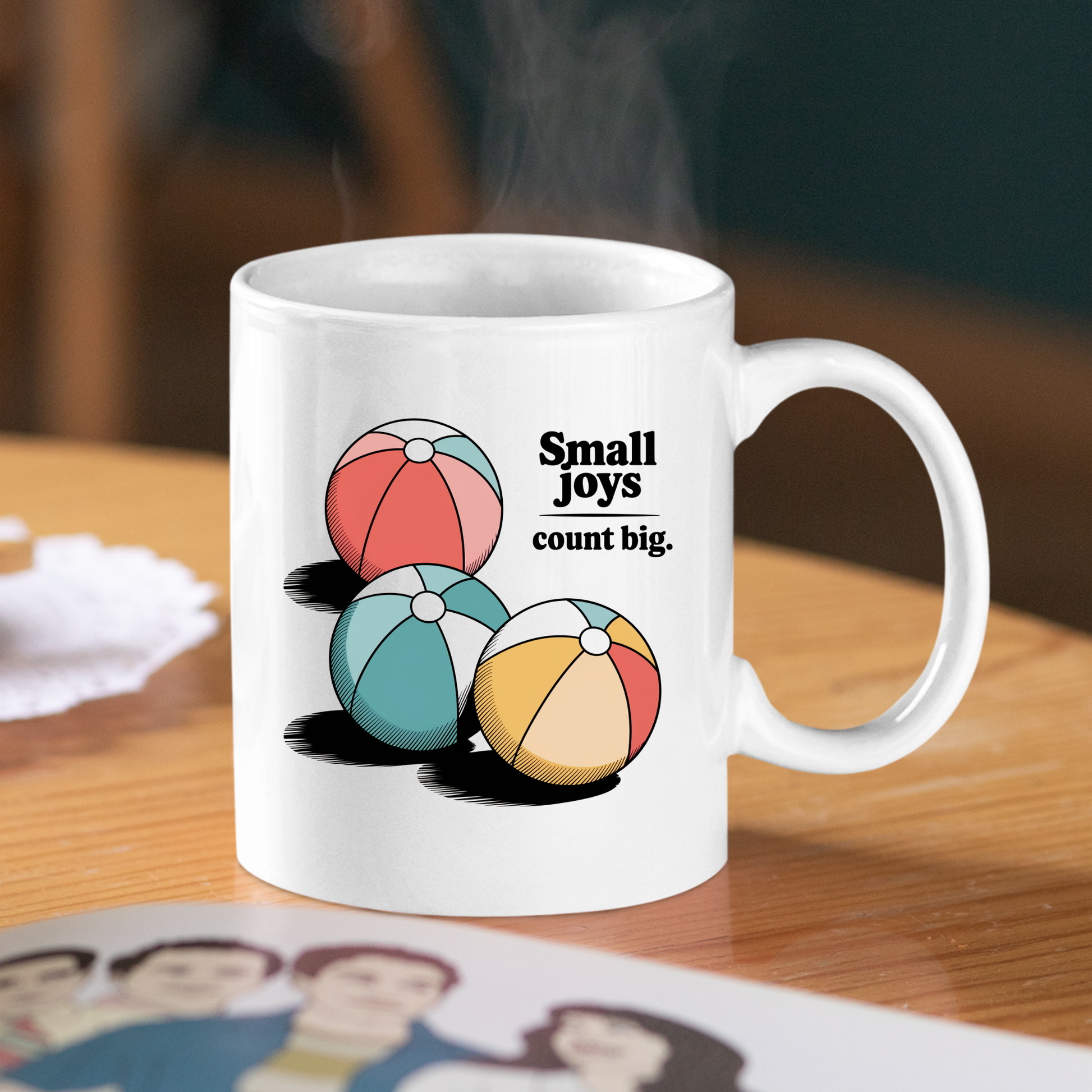 The Small Joys Count Big Mug | Inspirational Coffee Mug features a white ceramic design with three colorful retro-style beach balls and the uplifting phrase "Small joys count big." printed on it. Sitting on a wooden table, steam rises from this inspirational mug.
