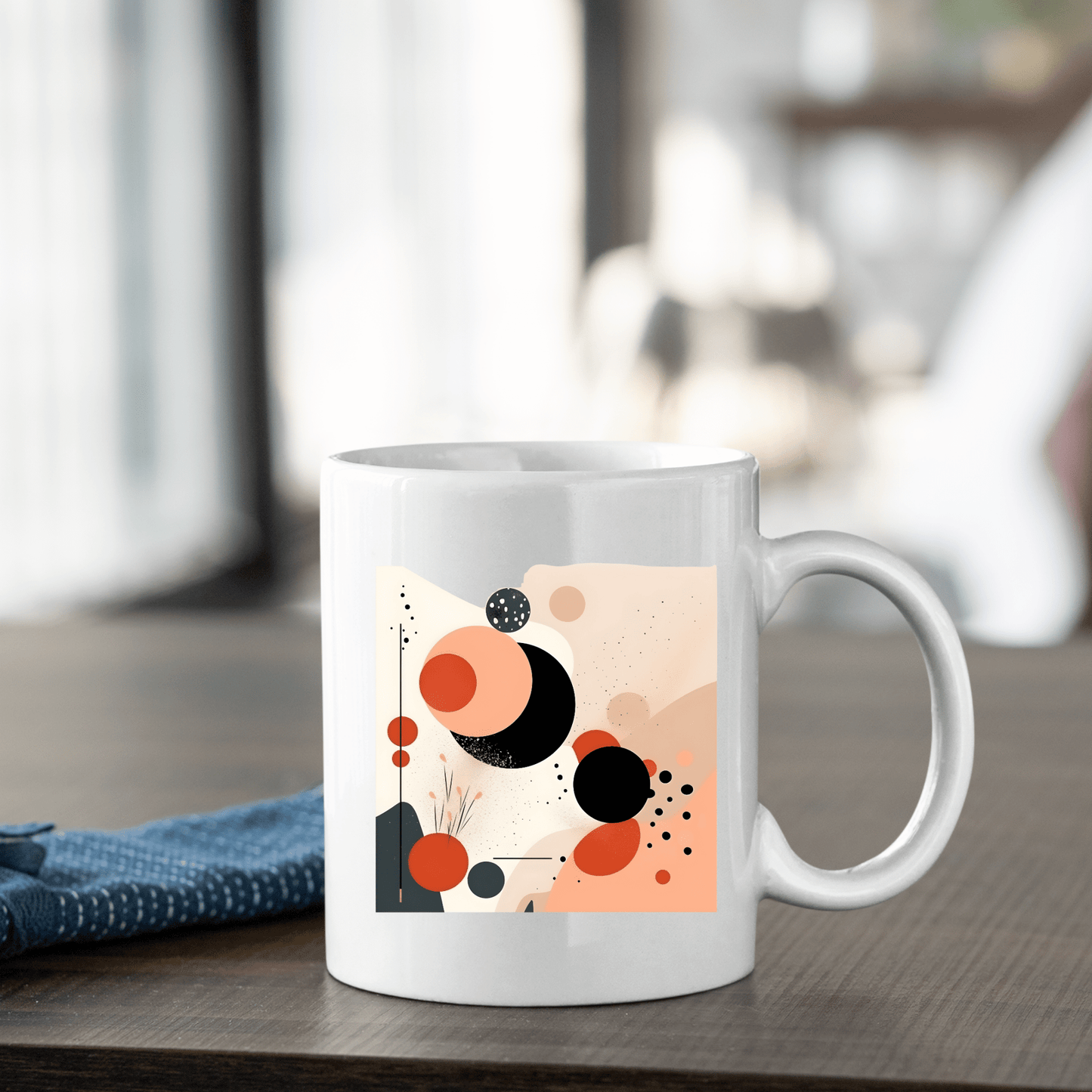 The Solar Bloom Mid Century Modern Mug, featuring an abstract geometric design in black, red, and beige, rests on a wooden table beside a folded blue cloth with a softly blurred background.