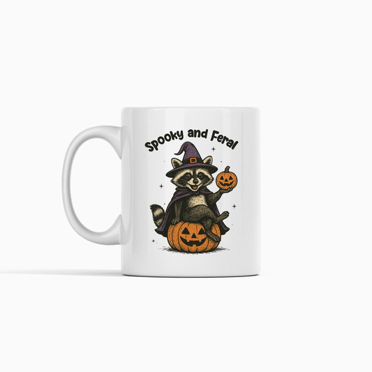 The Spooky and Feral Raccoon Mug is a white Halloween mug with a raccoon in a purple witch hat holding a jack-o'-lantern, perched on a pumpkin, with "Spooky and Feral" above—perfect as a funny Halloween gift!.