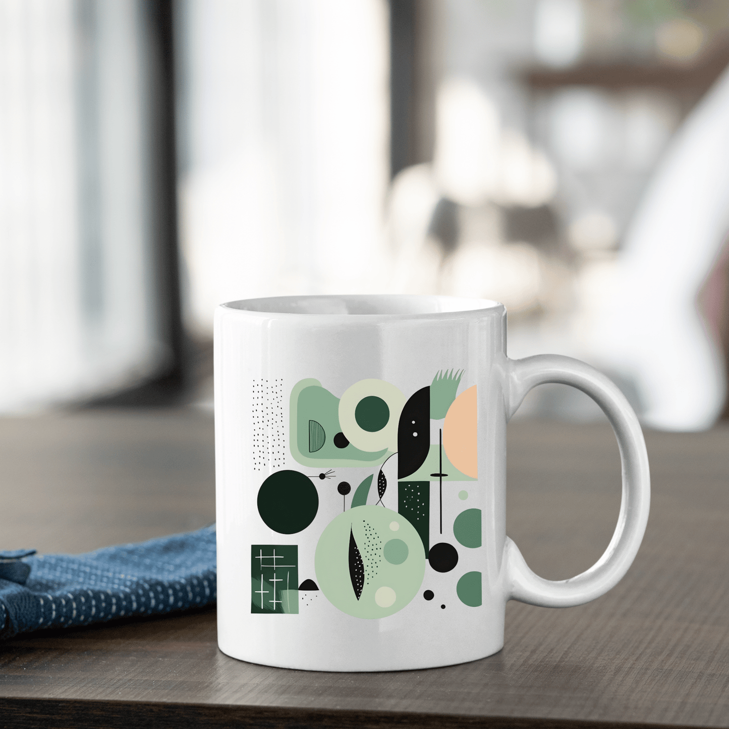 The Spring Geometry Mid Century Modern Mug, crafted from white ceramic and adorned with a green and black abstract design, sits on a wooden table beside a folded blue cloth against softly blurred nature-inspired décor.